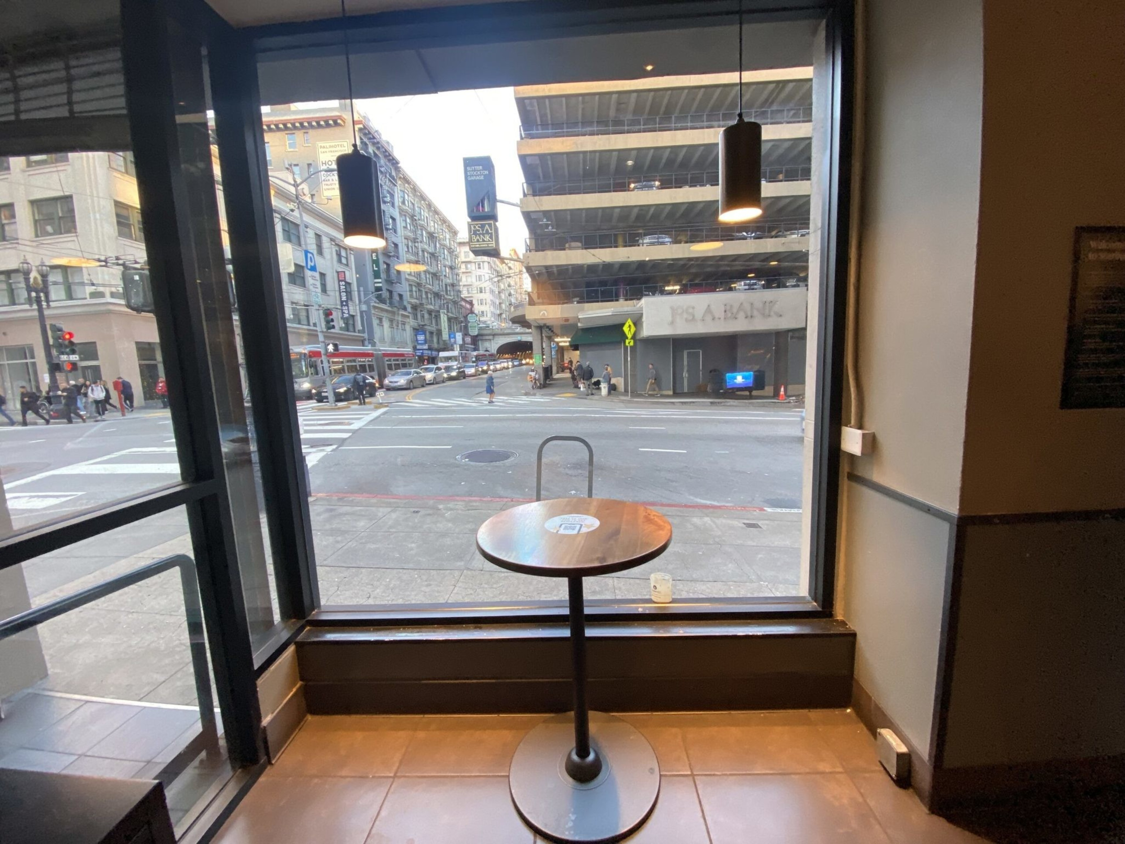 A small round table with a single chair is set by a large window overlooking a city street, traffic, pedestrians, and a multi-story parking garage.