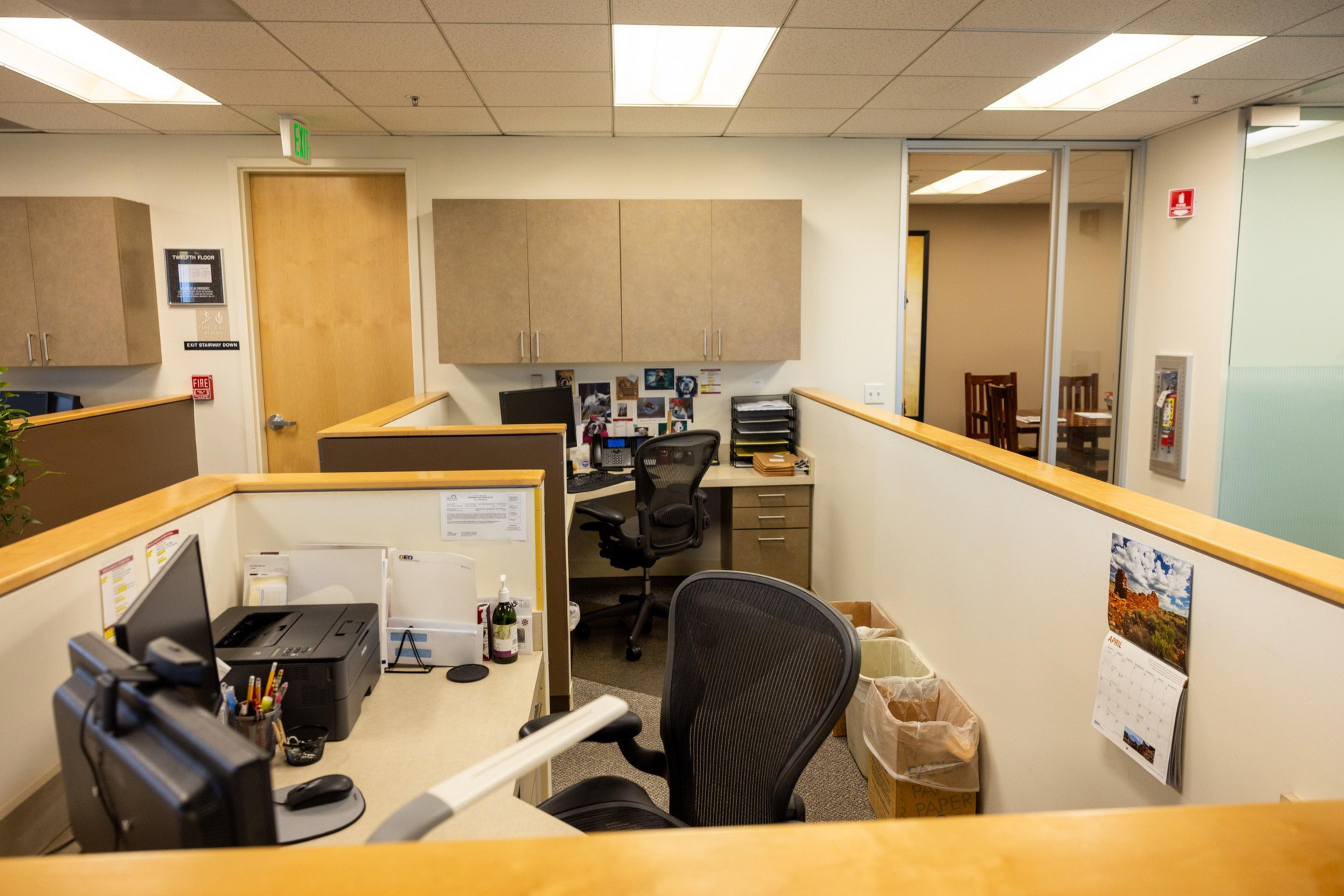 The image shows a tidy office cubicle with desks, chairs, computers, and cabinets. There are papers, stationery, and personal items on the desks, and a door, exit sign, and a calendar.