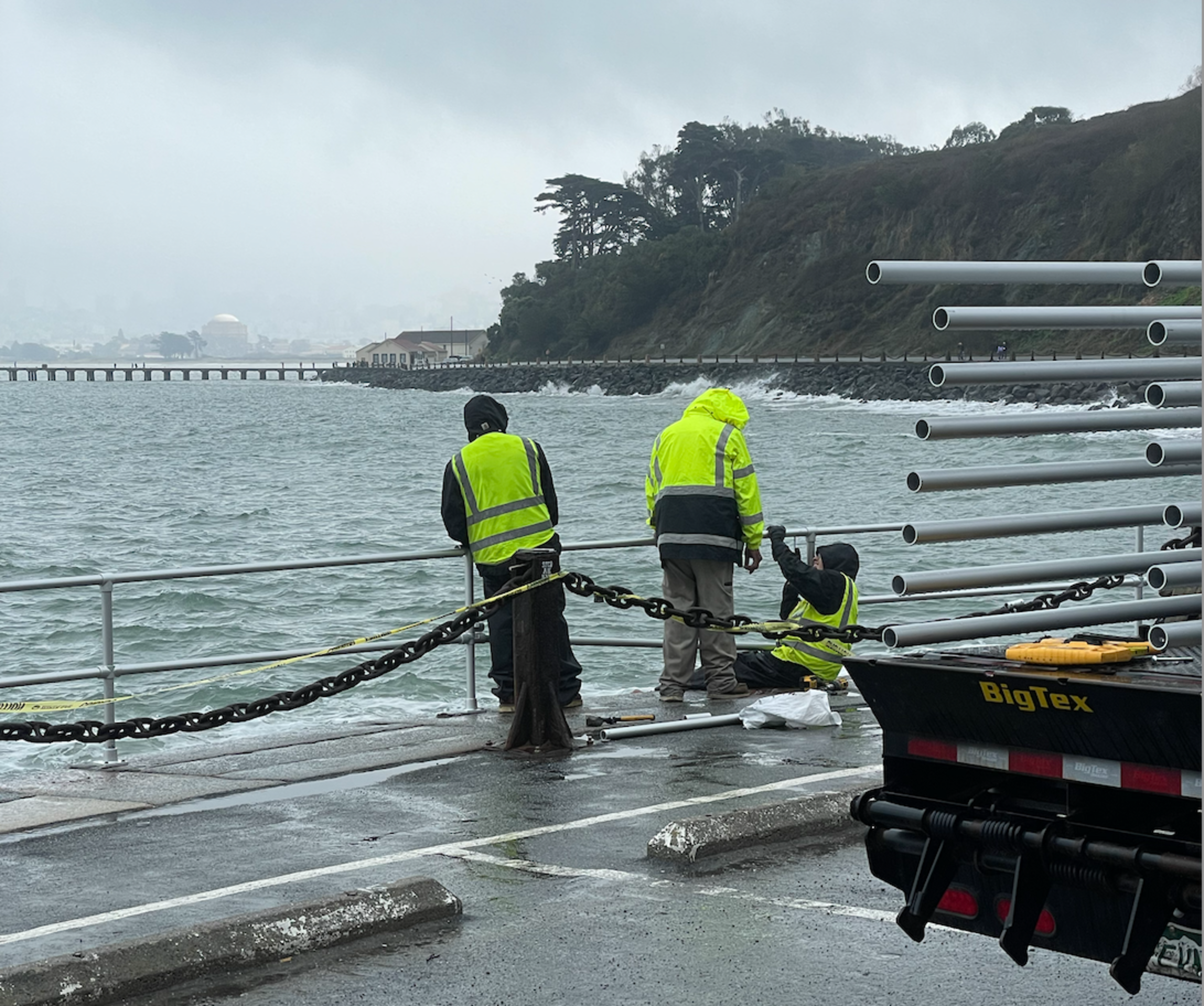 Three people in fluorescent green vests and hooded jackets stand over a low metal fence by a large body of water.