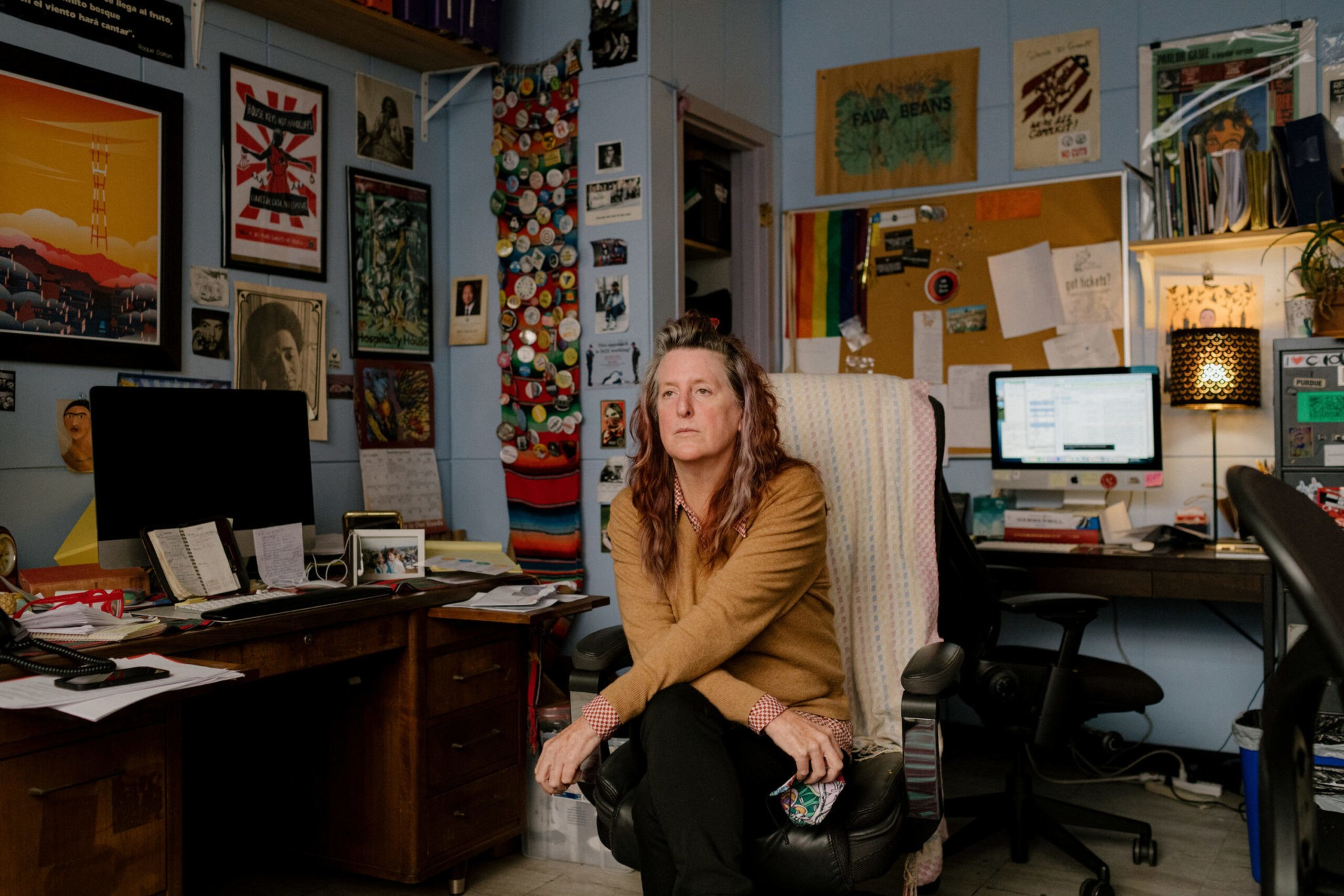 A person sits in a chair in a cluttered office filled with colorful posters, a computer, books, and a wall covered in badges and notes. They look thoughtful.