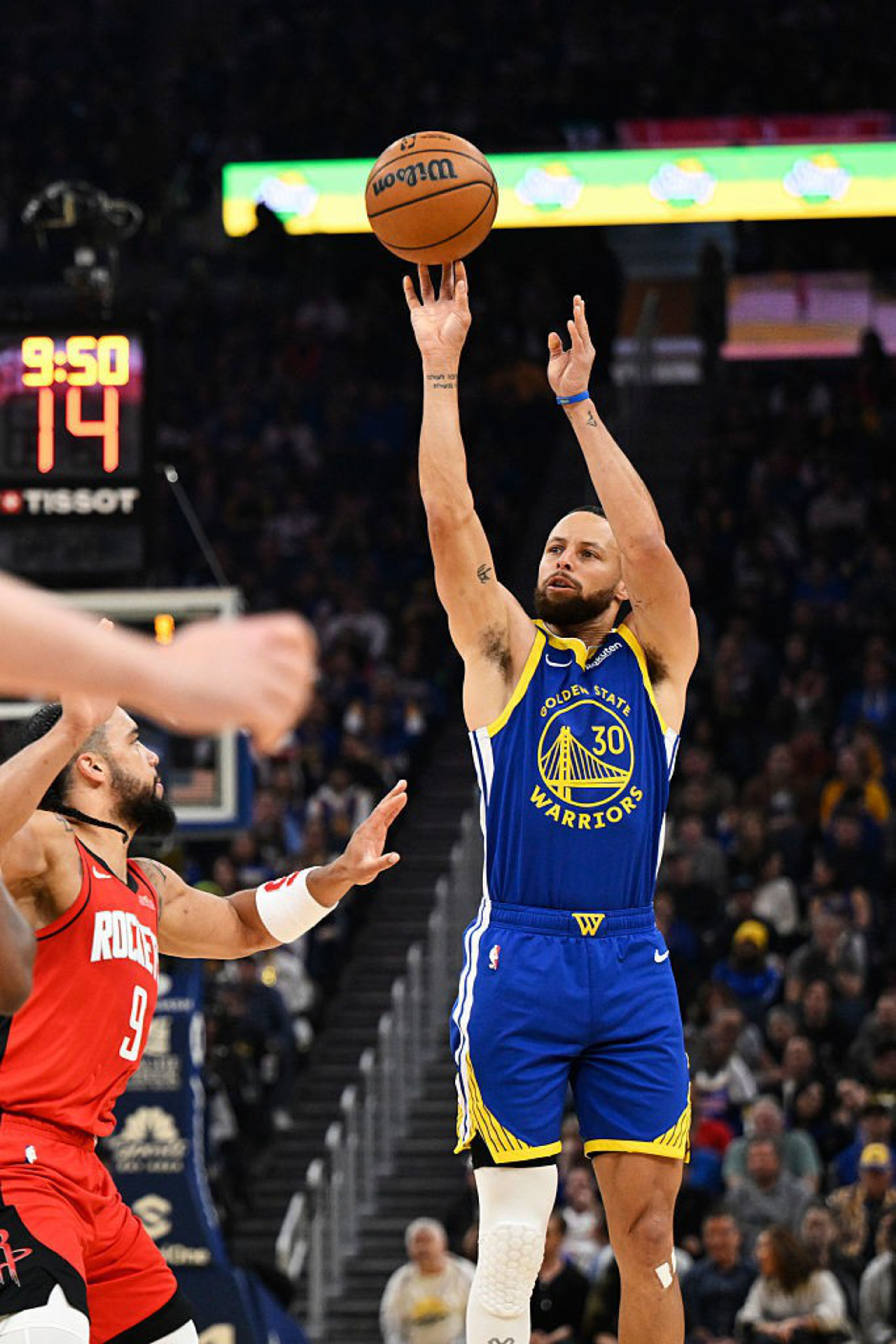A basketball player in a blue Golden State Warriors jersey is shooting the ball mid-air. Another player in a red Rockets jersey is nearby. The scoreboard shows 9:50.