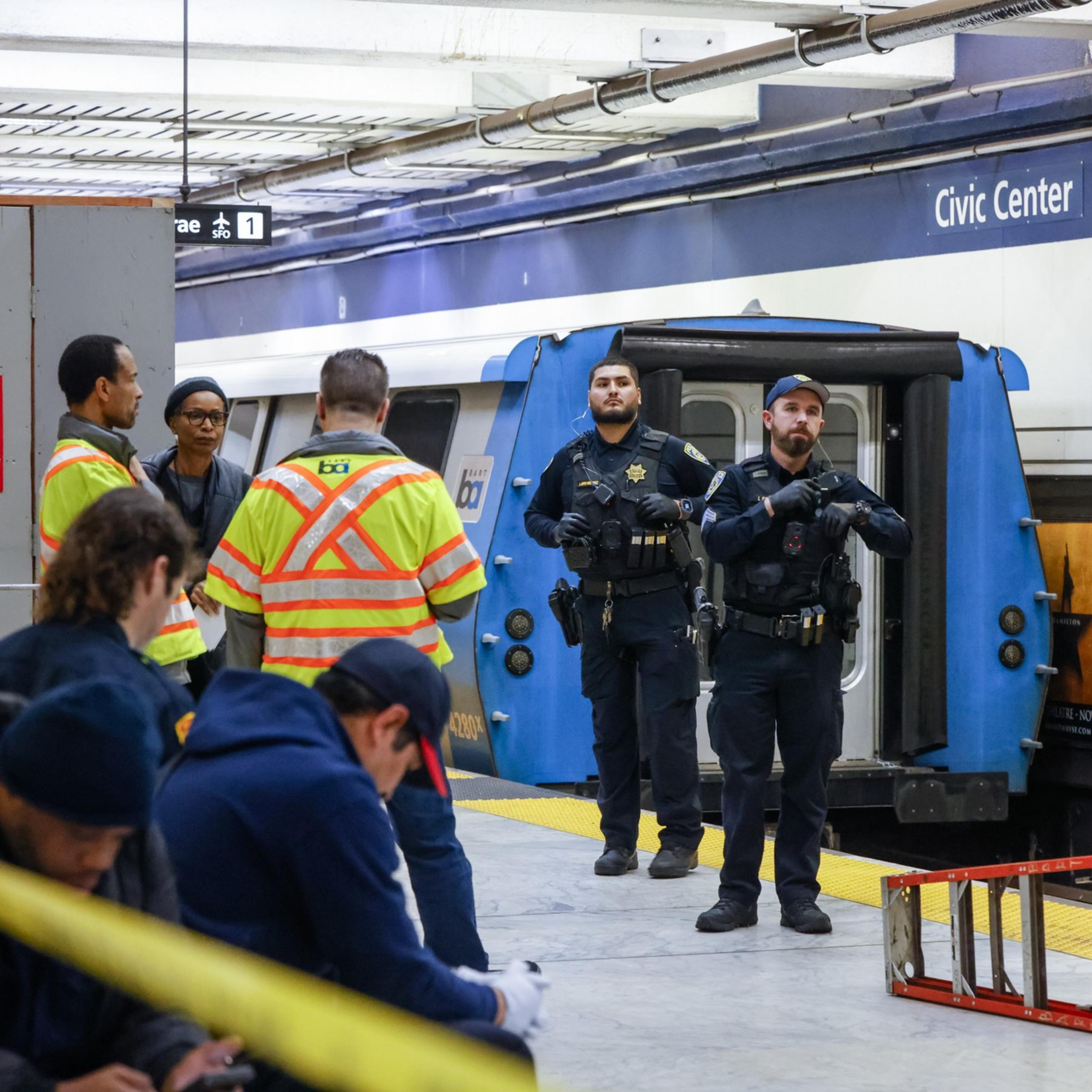 Two police officers stand near a train at Civic Center station. Workers in high-visibility jackets converse nearby, with a few people seated in the foreground.
