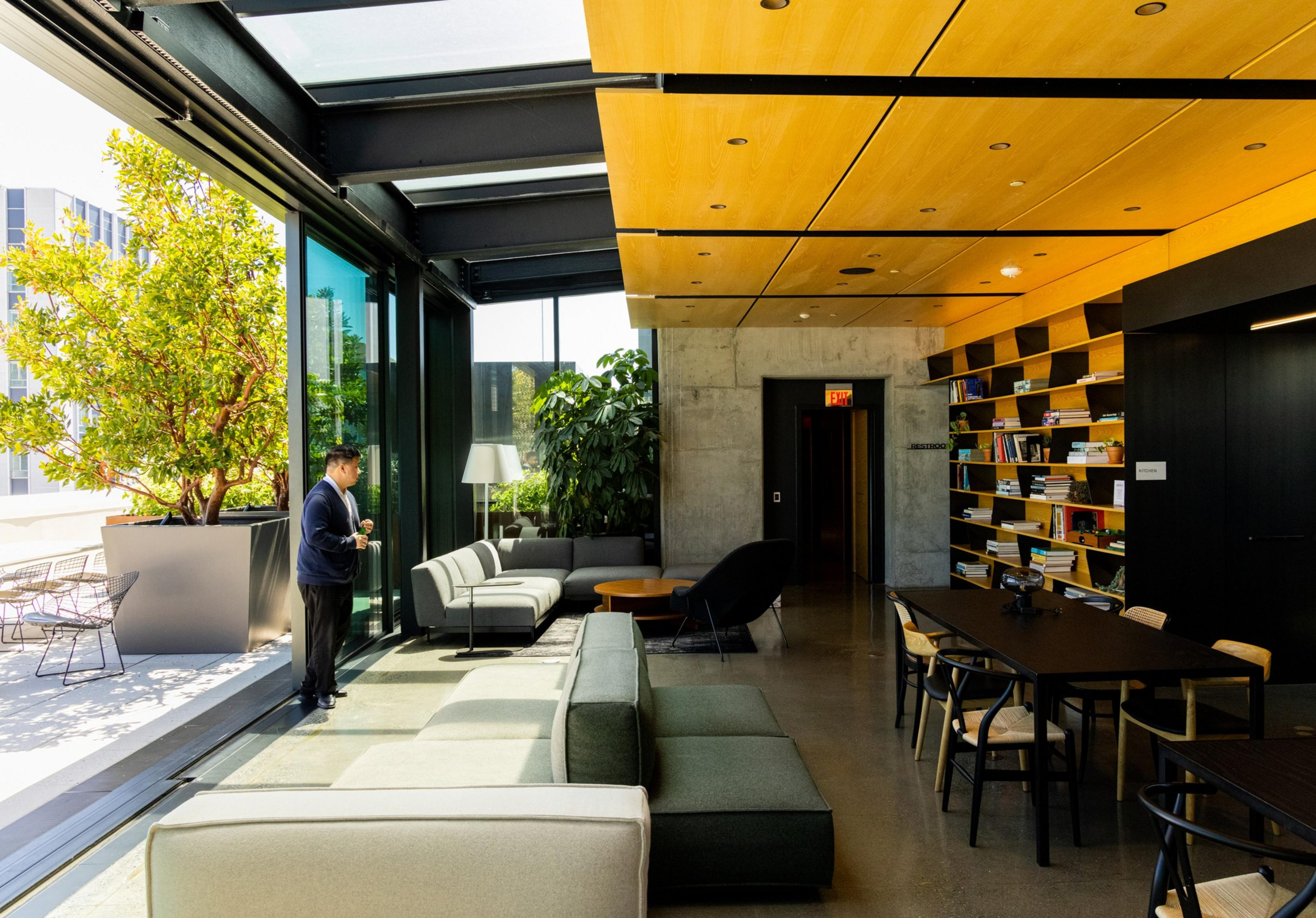 A modern room with large windows, couches, a person standing, bookshelves, a table with chairs, and some greenery inside and outside. The room has an outdoor view.