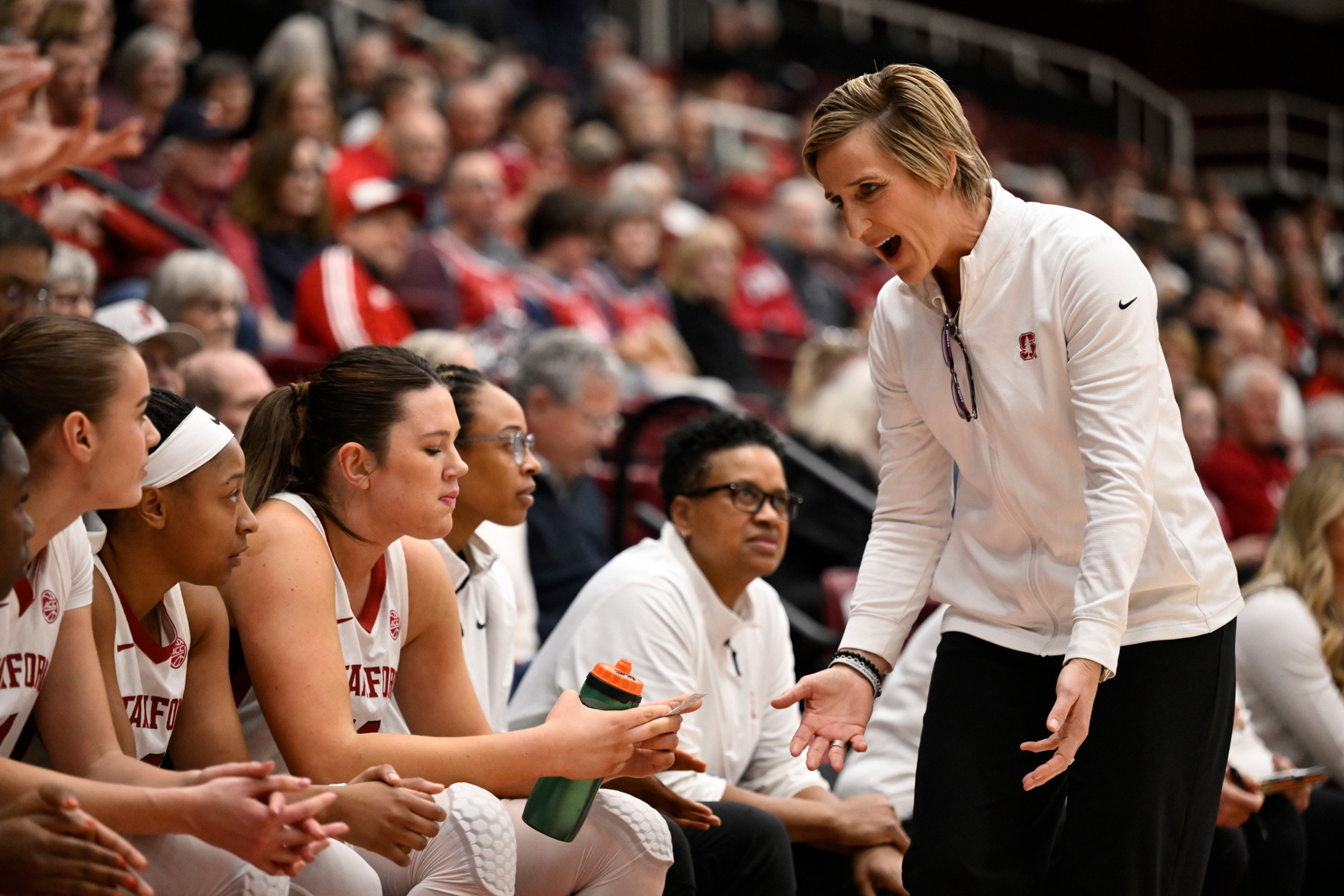 A basketball coach in white talks passionately to seated Stanford players during a game timeout, with a focused crowd in the background.
