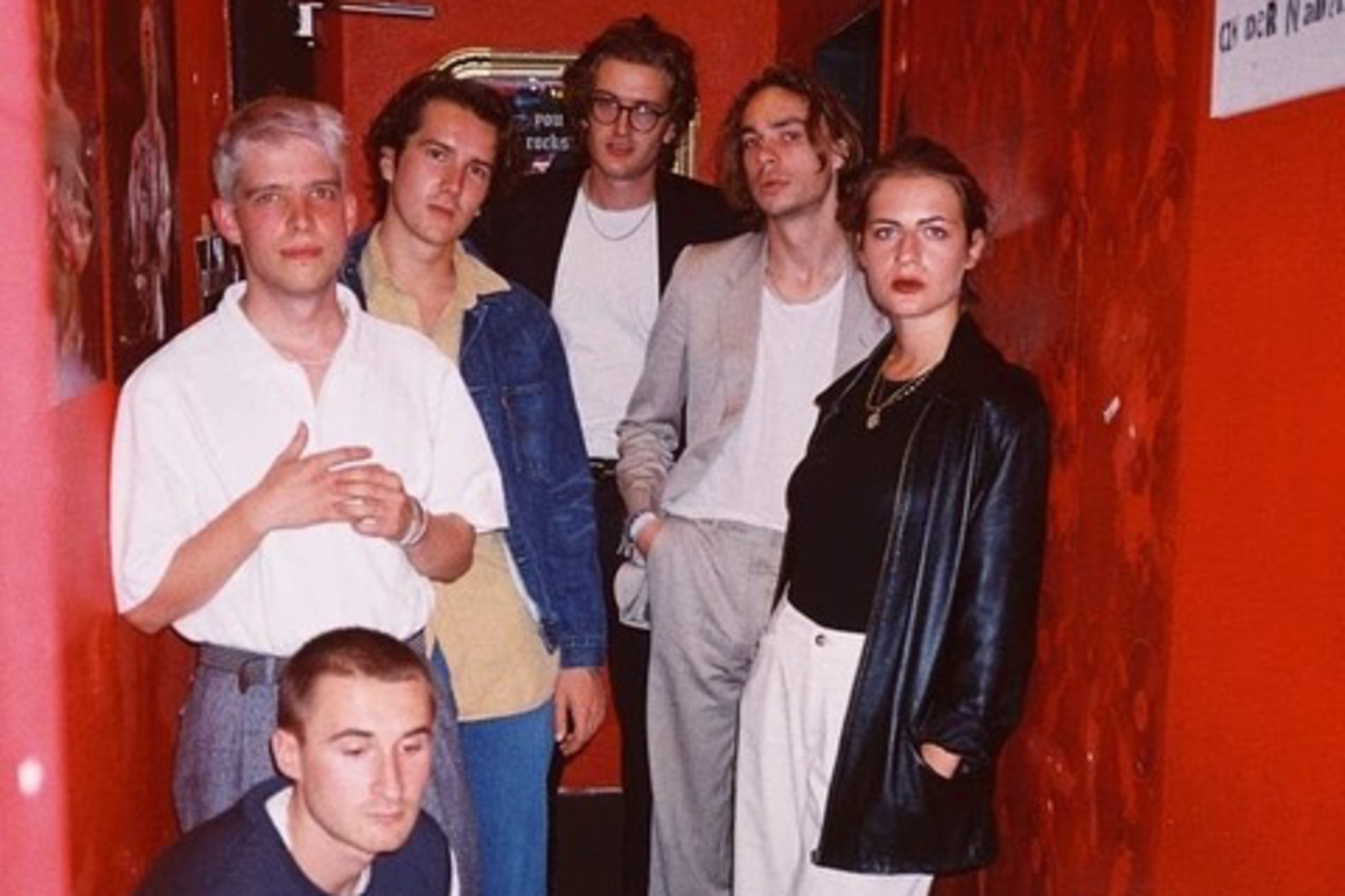 Six people are casually posed in a narrow, red-walled hallway. They wear a mix of casual and smart clothing, and their expressions are mostly relaxed.