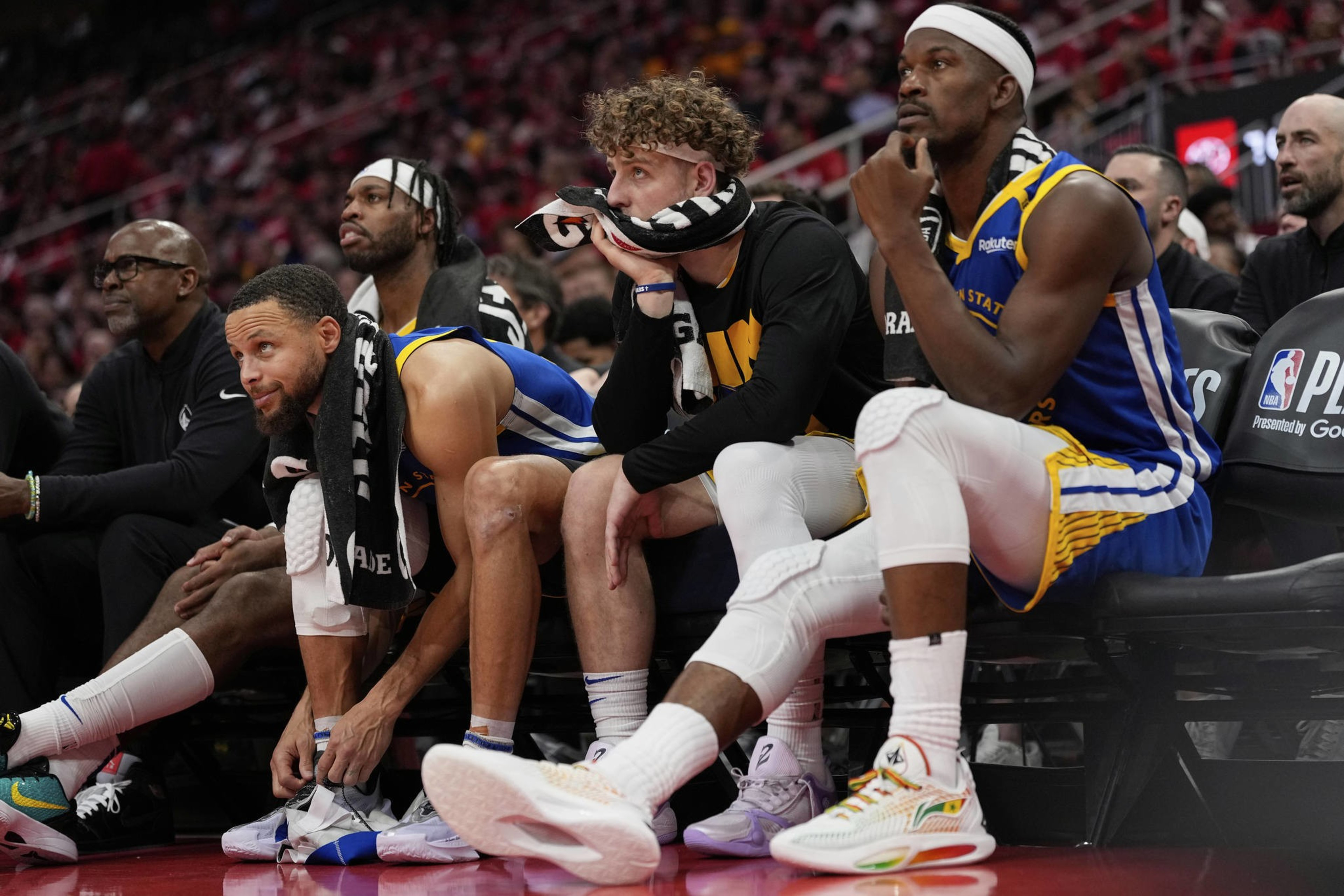 Four basketball players sit on a bench, looking pensive. Two wear blue and yellow jerseys while the others have black and yellow. Towels are draped around their necks.