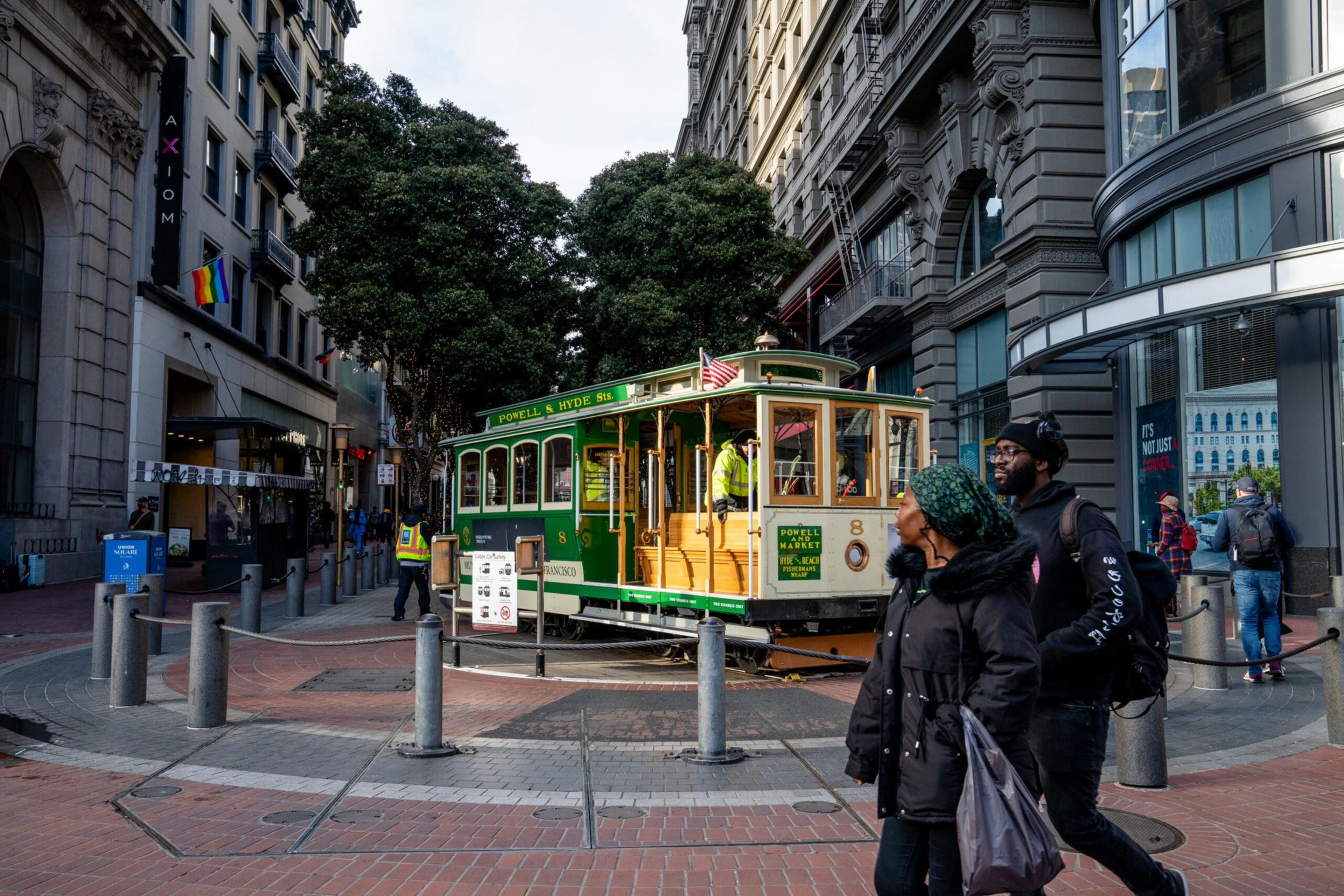 A green and cream cable car labeled "Powell & Hyde St" is on a turntable surrounded by tall buildings, trees, and pedestrians walking nearby.