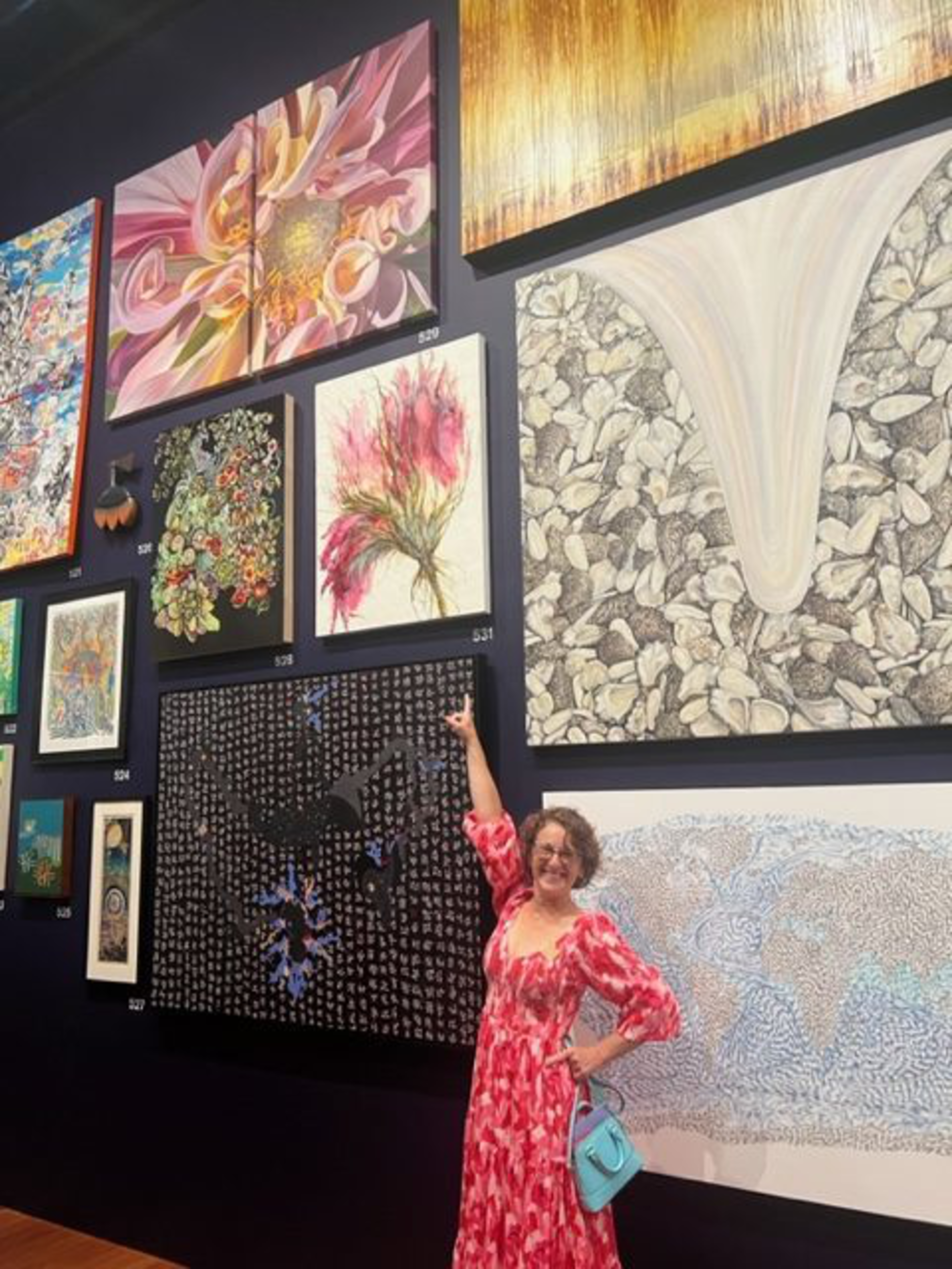 A woman in a red and white dress points to a pink flower painting on a wall filled with various colorful and abstract artworks.