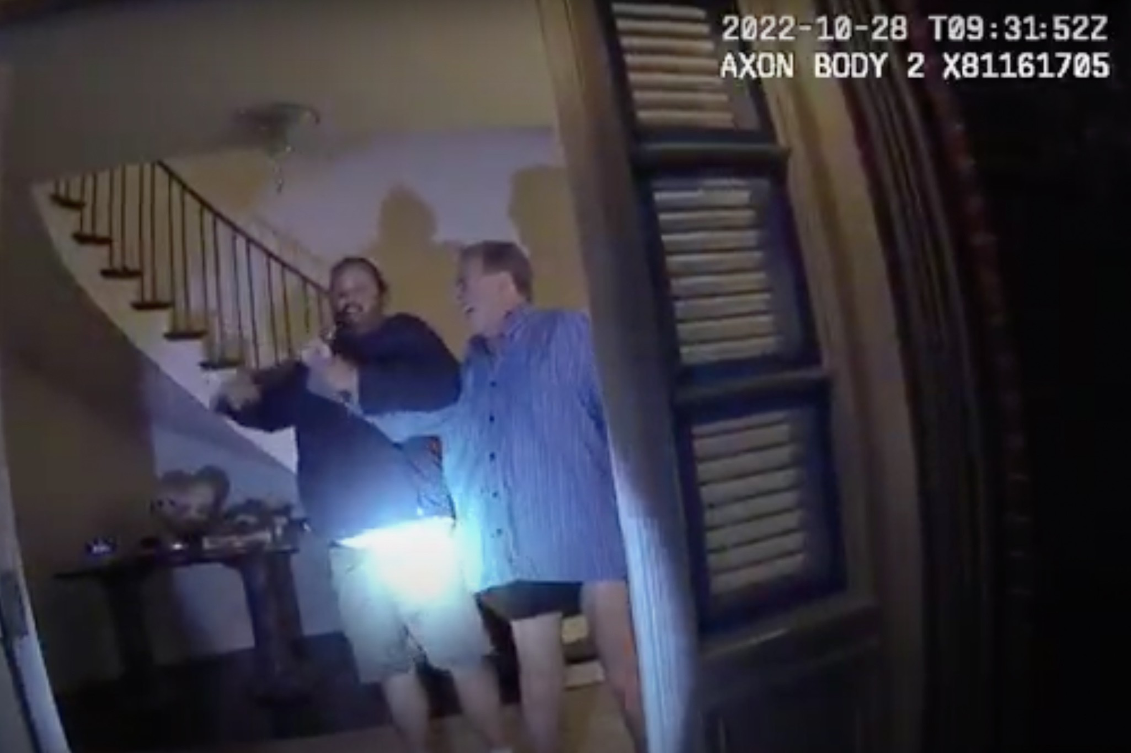 Two men stand inside an open doorway; one holds a flashlight illuminating the scene. A staircase is visible in the background.