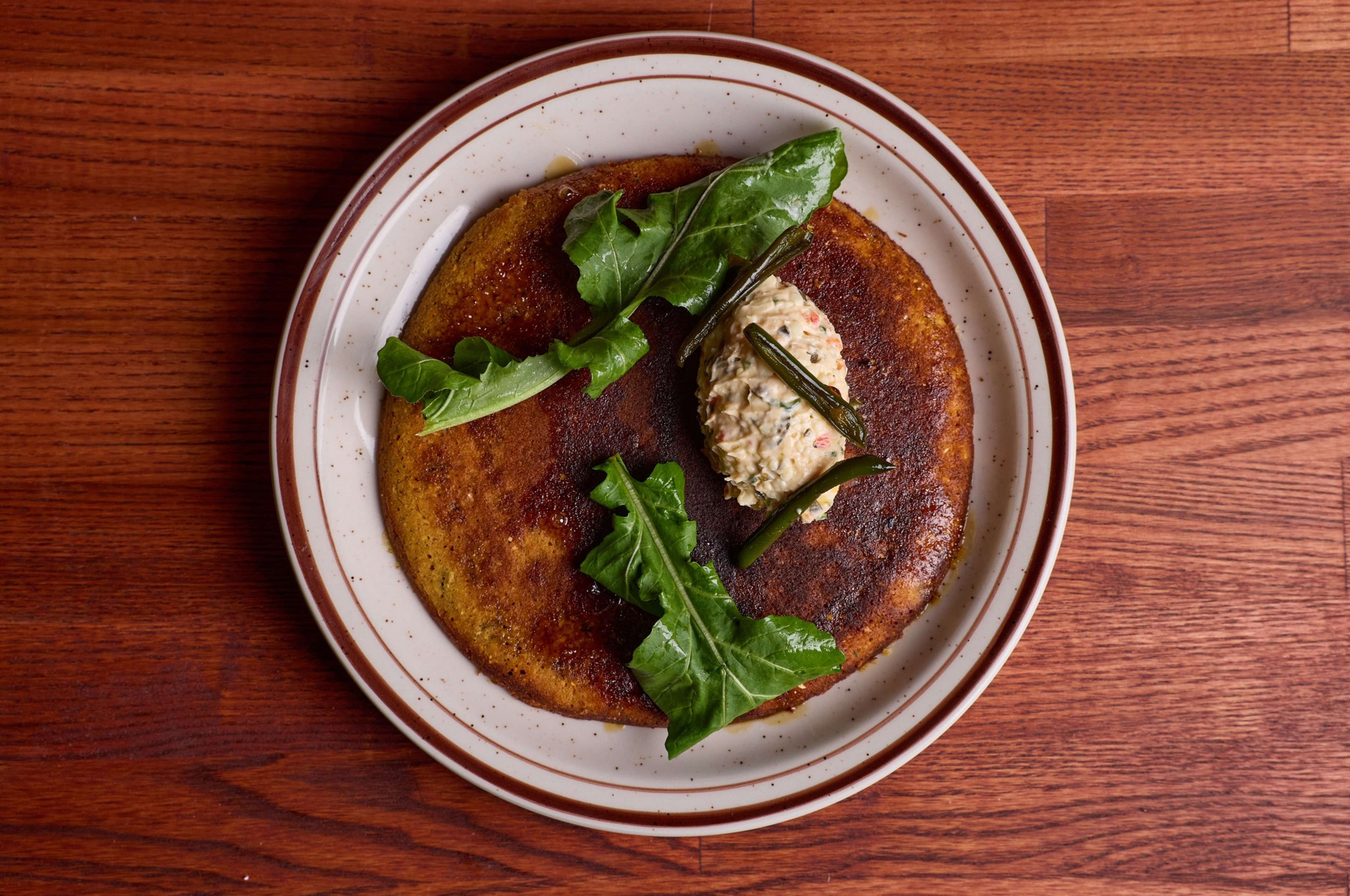 A browned pancake topped with a dollop of creamy spread, dark green leafy greens, and thin green garnish sits on a white plate with brown trim.