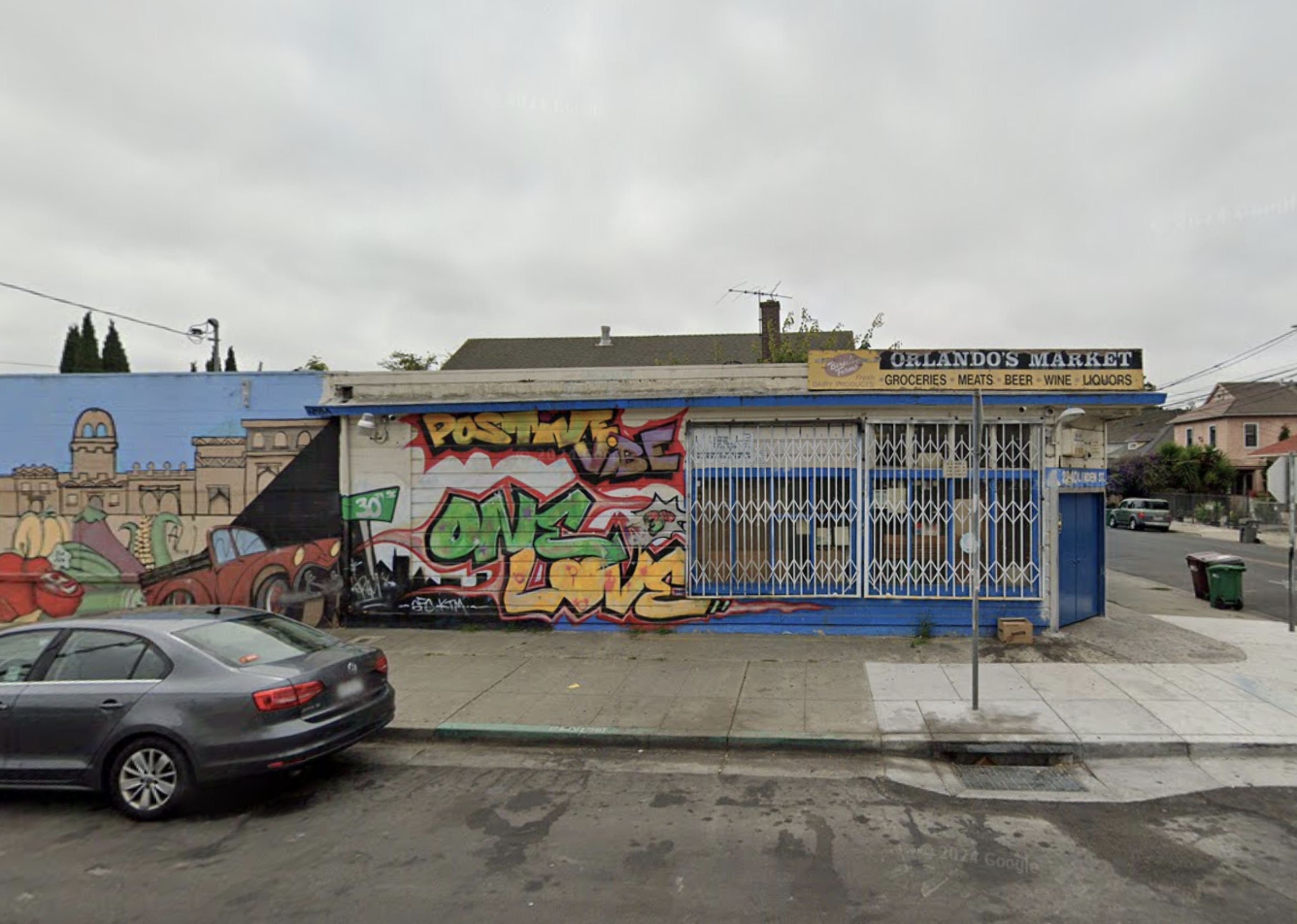 A small market with a colorful graffiti wall stands closed behind a metal gate, on a street with parked cars.