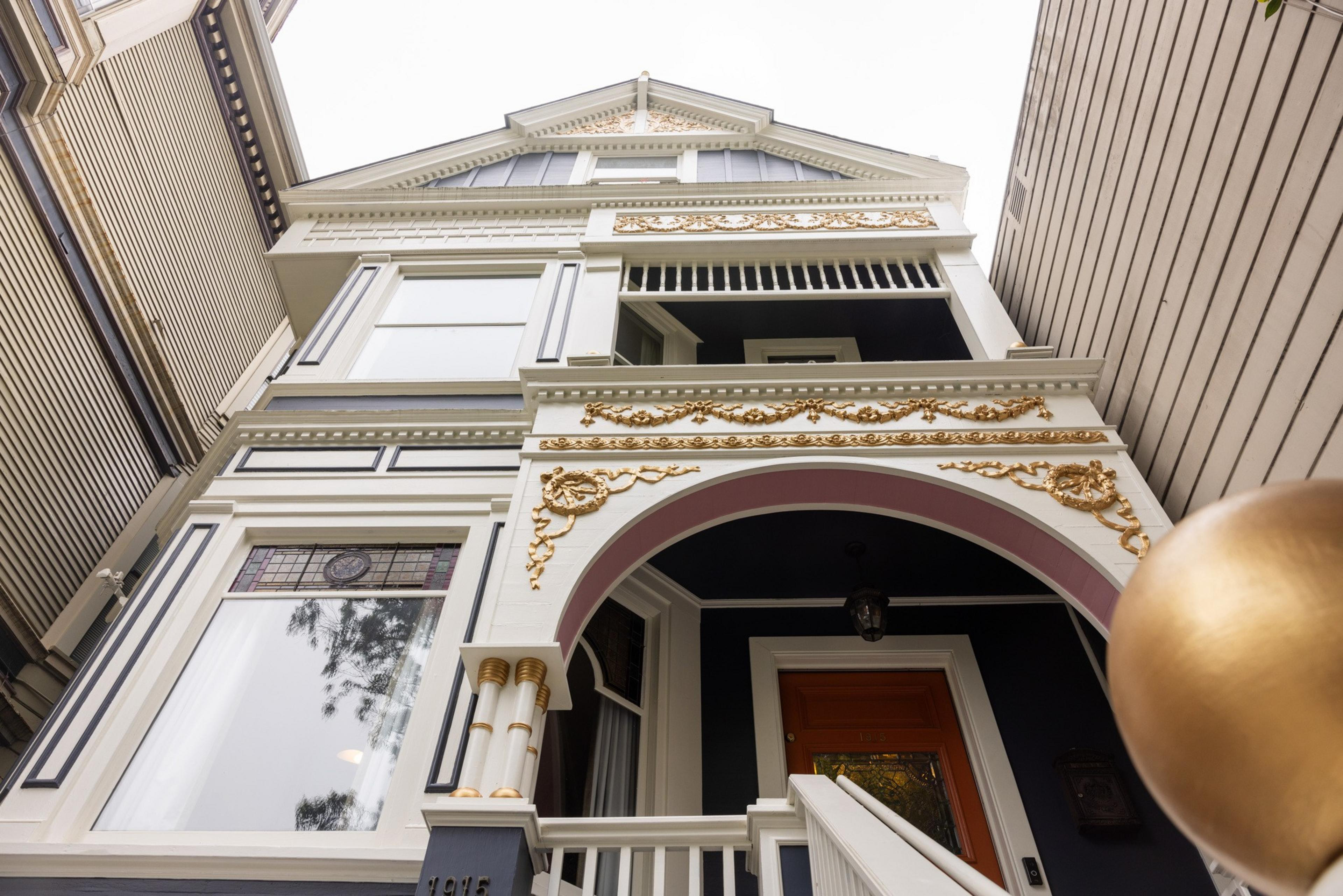 The image depicts a multi-story Victorian house with ornate white and gold trim, stained glass windows, and a prominent red door.