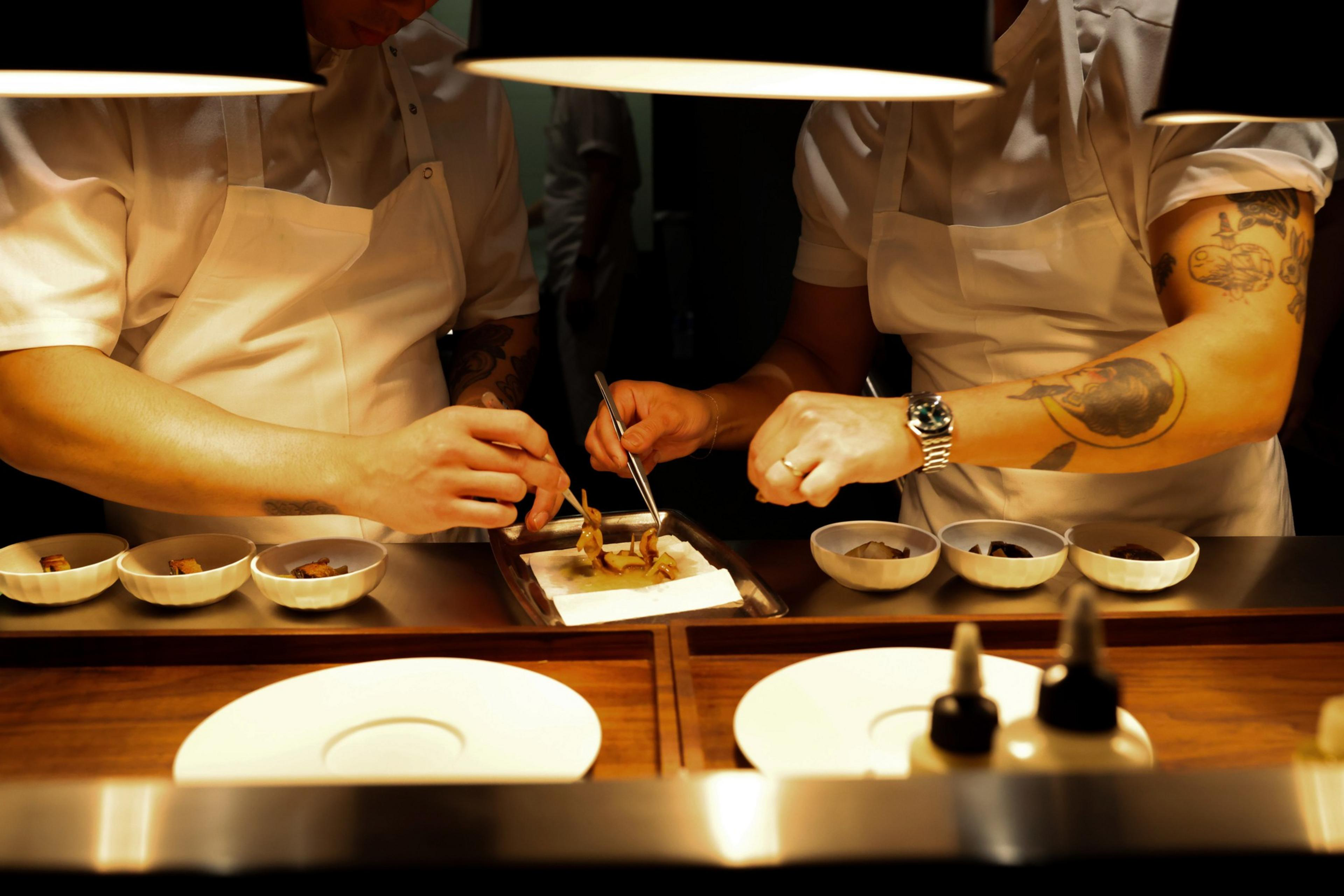 Two chefs in white uniforms and aprons meticulously plate a dish under warm lighting, surrounded by small bowls and sauces on a wooden countertop.
