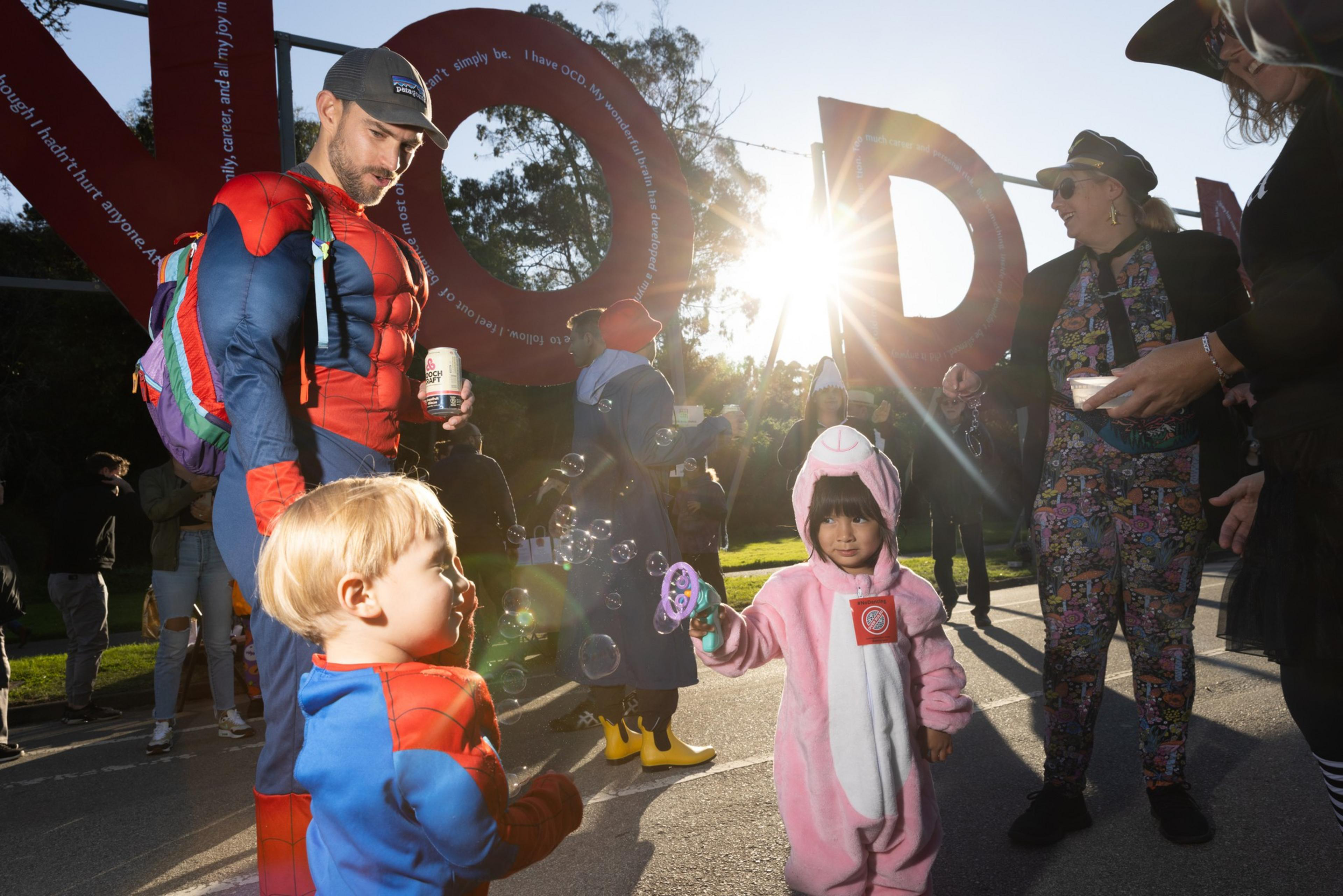 A man in a Spider-Man costume watches a young child in a similar outfit while another child in a pink animal costume blows bubbles, with adults nearby.