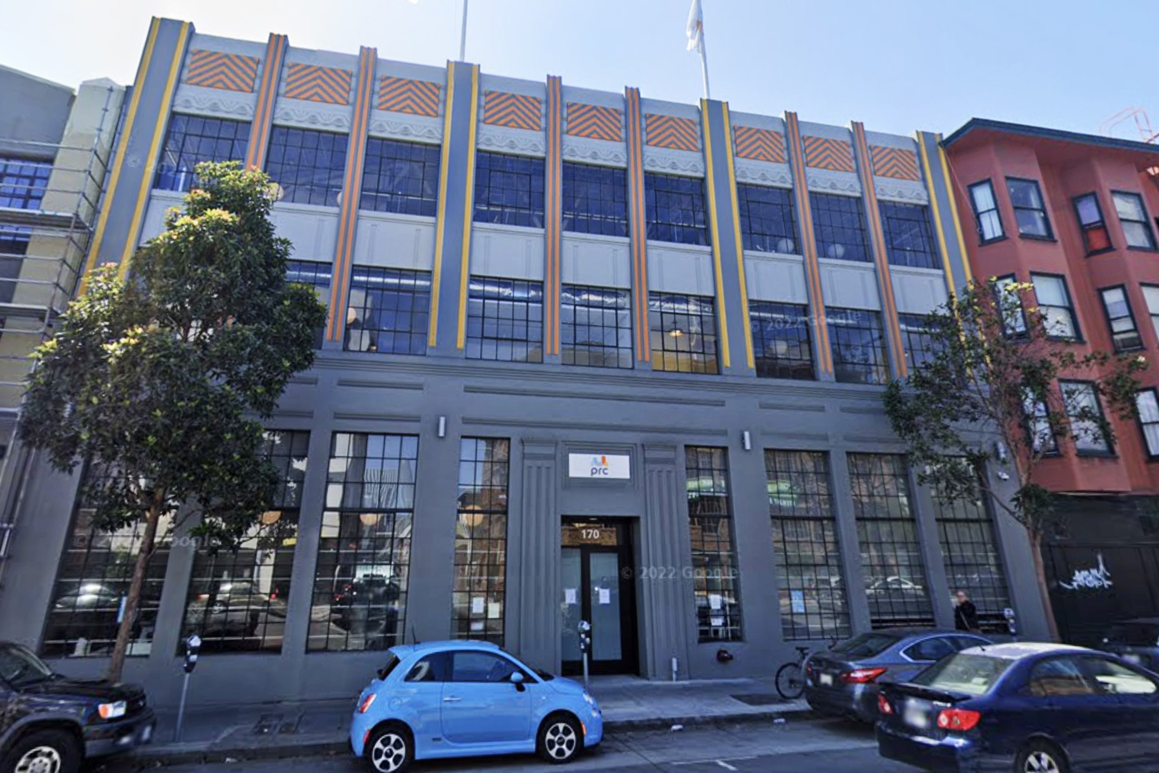 A three-story building with tall windows and decorative vertical yellow and orange stripes stands on a city street with parked cars and trees nearby.