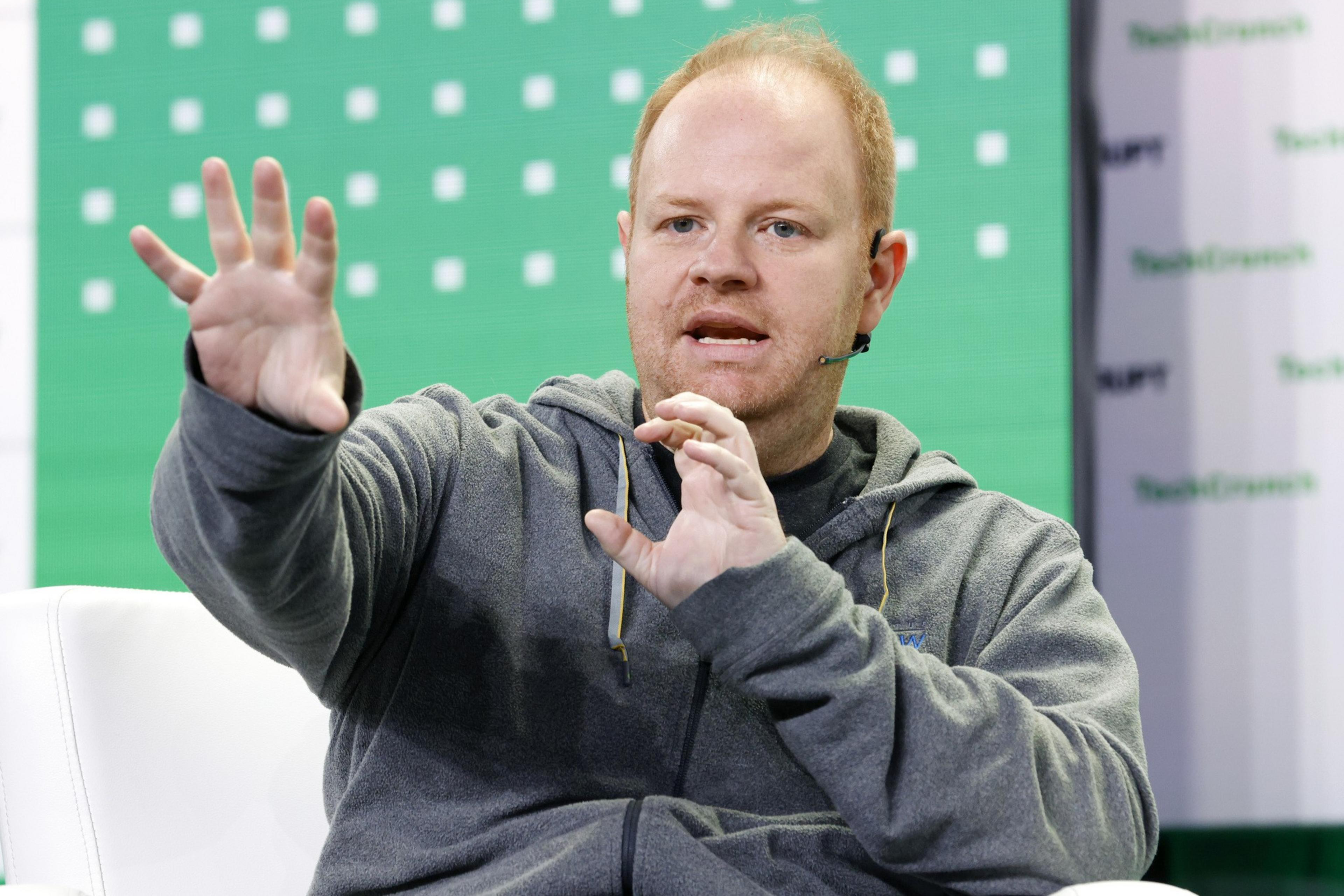 A person in a gray hoodie is speaking on stage with a microphone. The background is green with white squares and partially visible text.