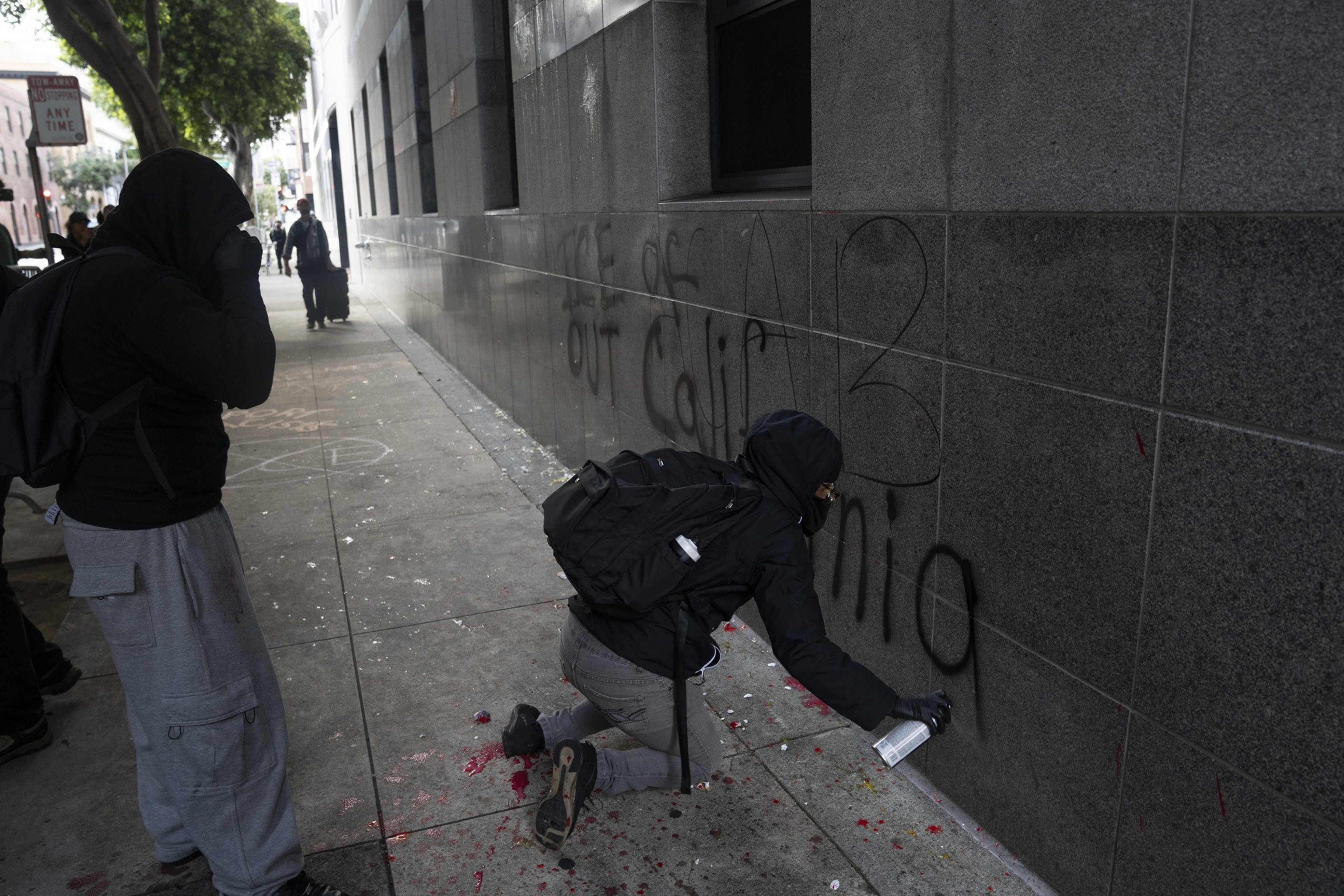 A person in dark clothing is spray-painting graffiti on a building's gray wall. Another person stands nearby, both partially obscured by hooded garments.