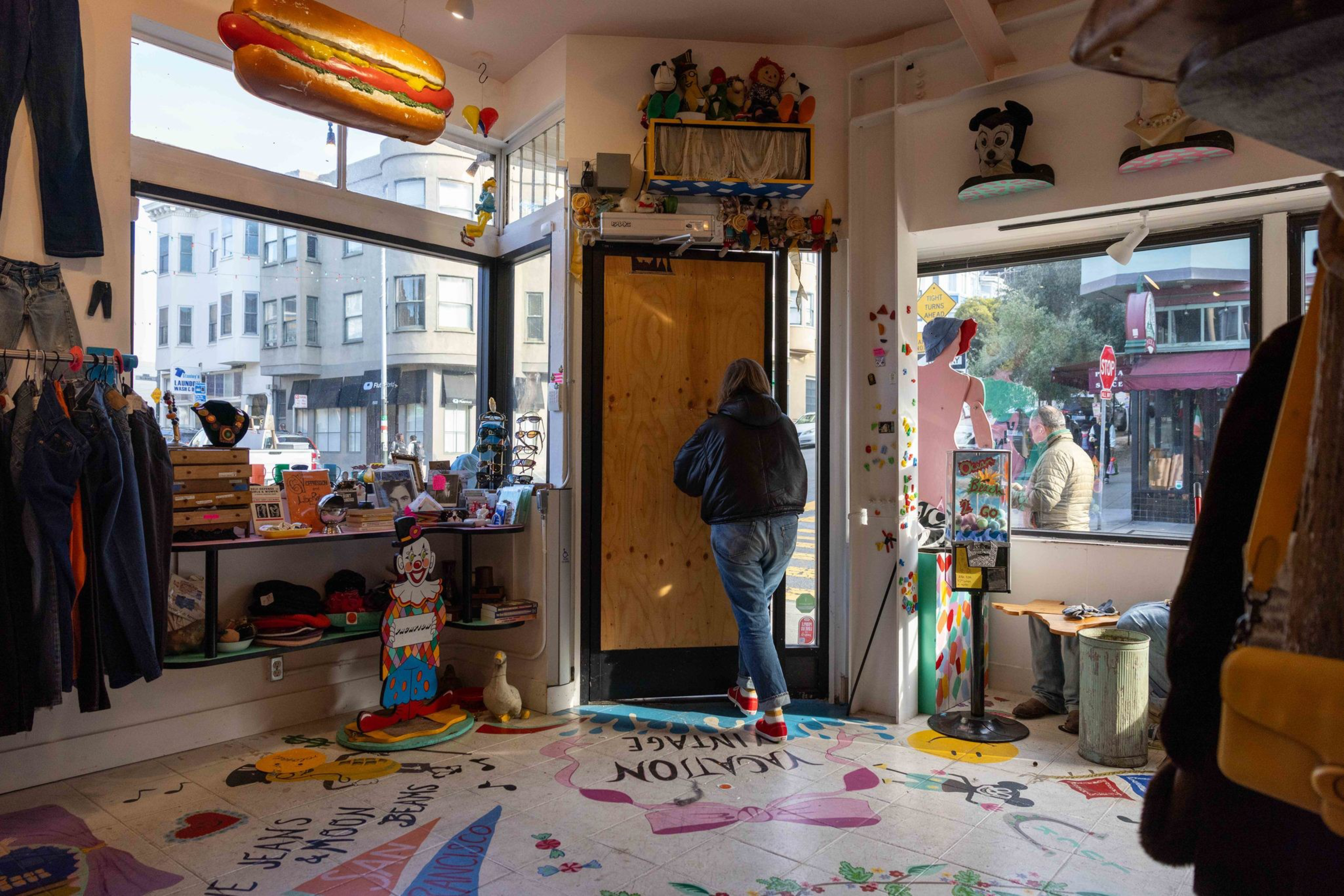 A person in a hooded jacket walks toward a wooden door inside a quirky store filled with colorful decor, clothing racks, and novelty items.