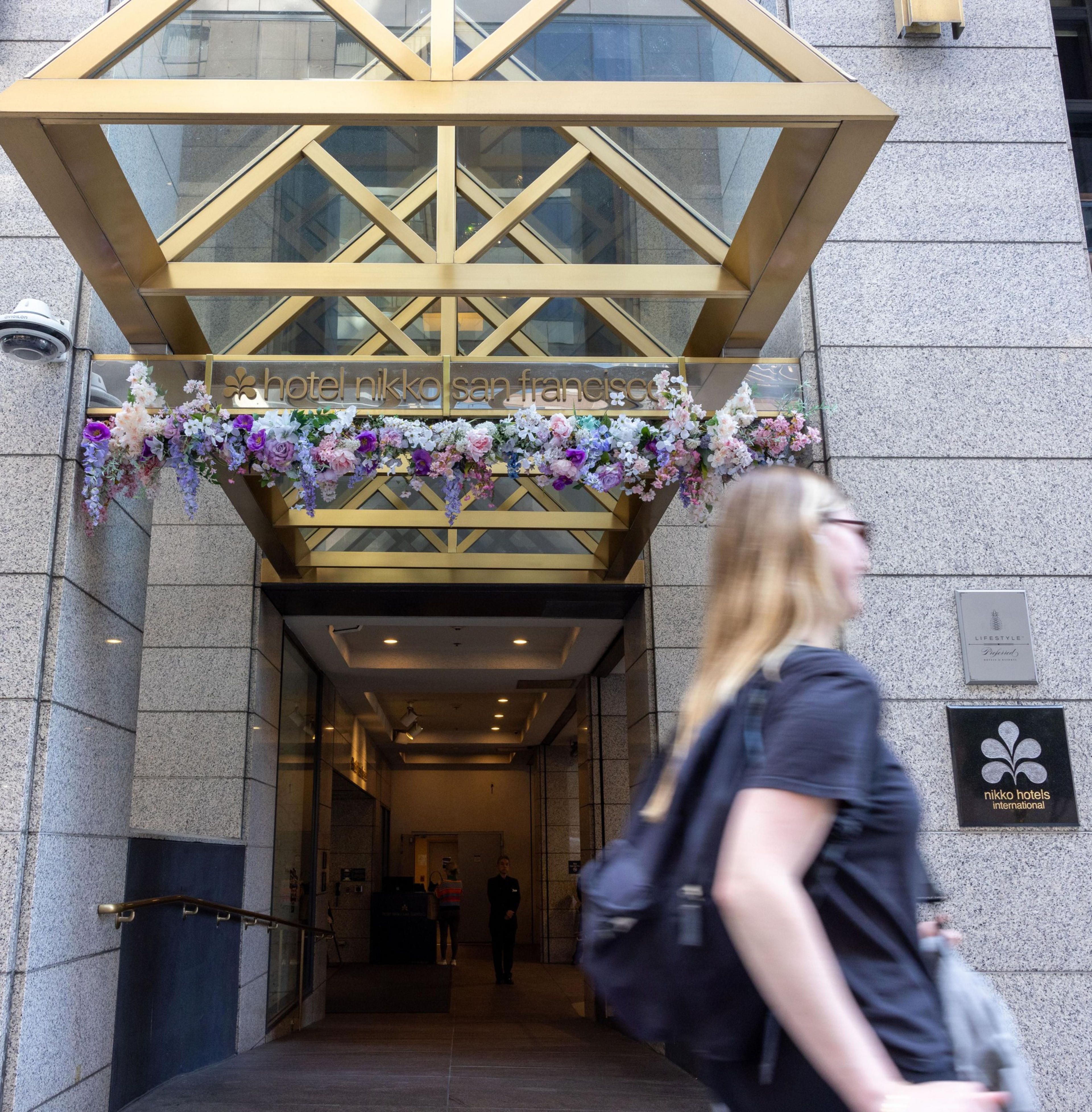 The image shows the entrance of Hotel Nikko San Francisco with a floral arrangement above the entrance. A person with a backpack walks by in the foreground.