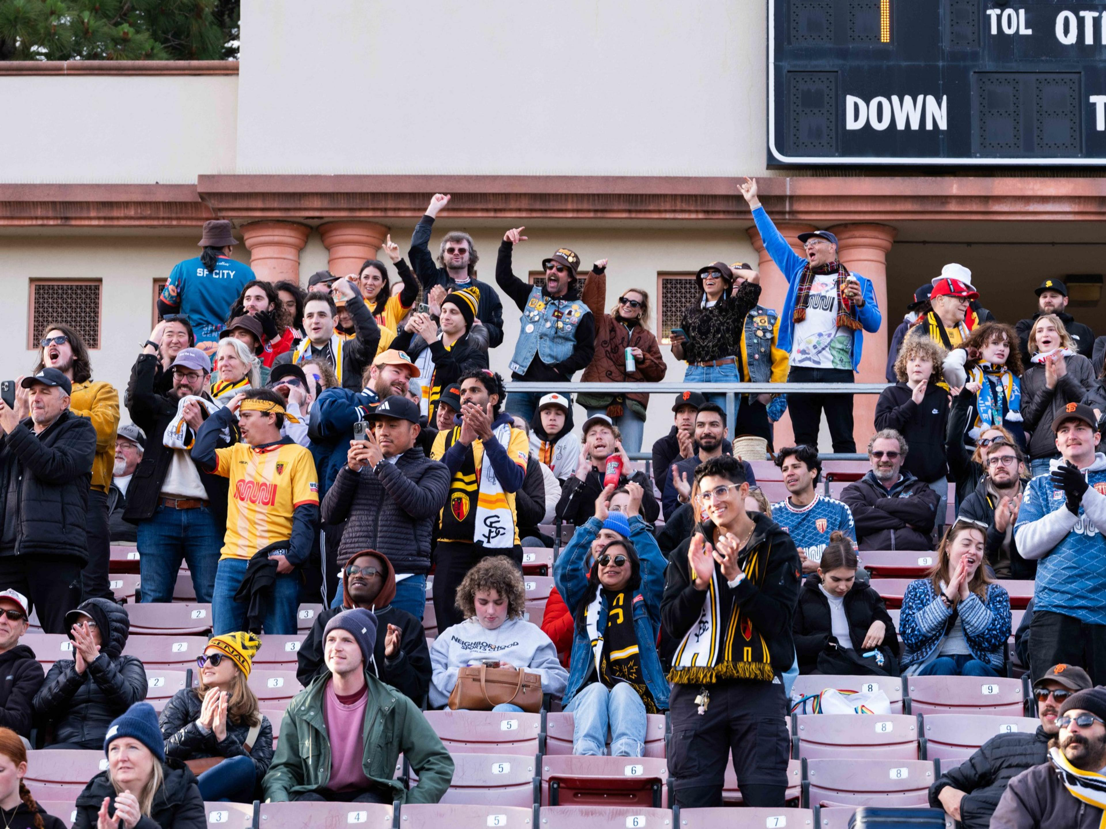The image shows a lively crowd in a stadium, many wearing sports jerseys and scarves, cheering and clapping enthusiastically. The scoreboard is visible above them.