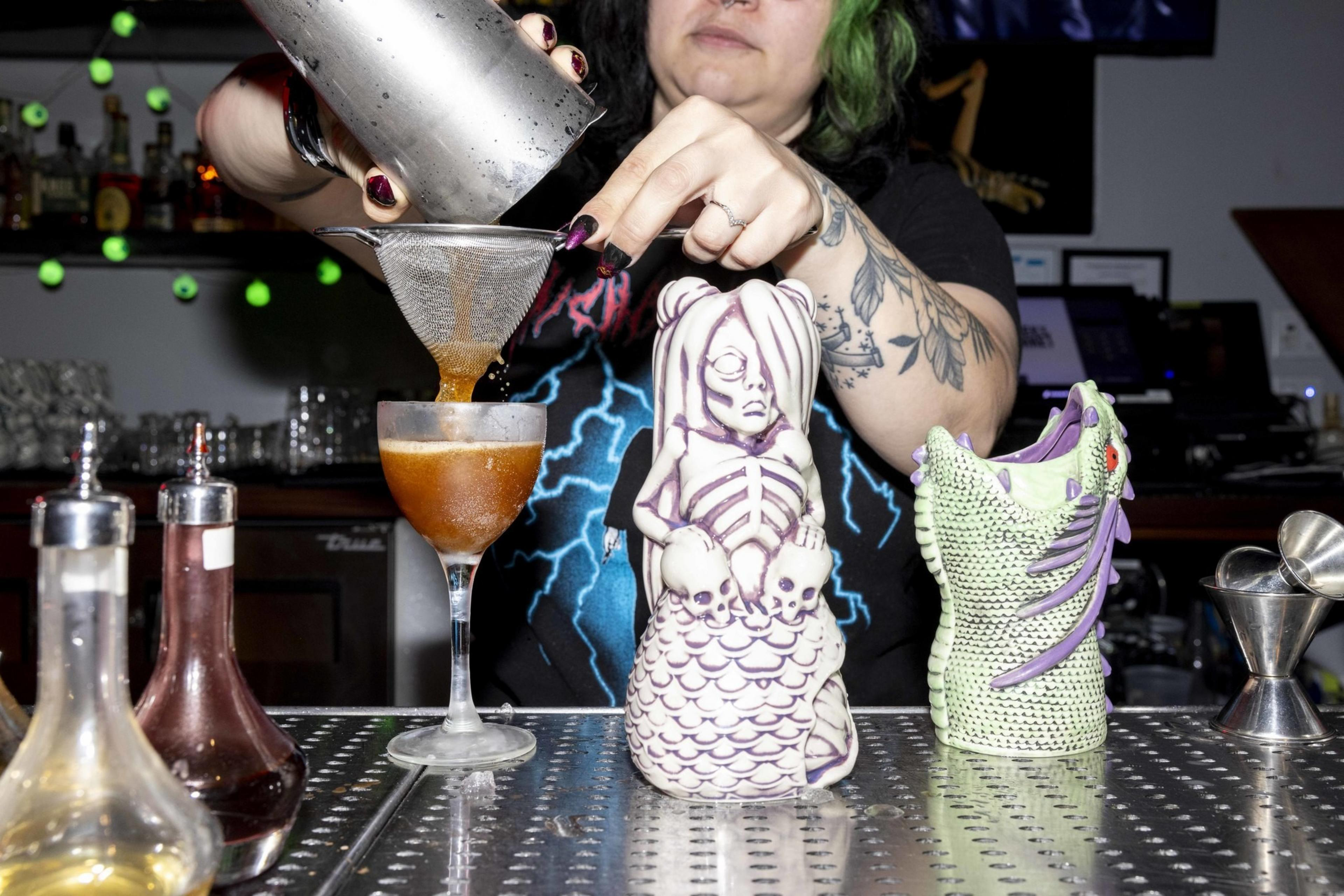 A person with tattoos is straining a cocktail into a glass at a bar, with two decorative ceramic mugsâ€”a mermaid and a monsterâ€”on the counter.
