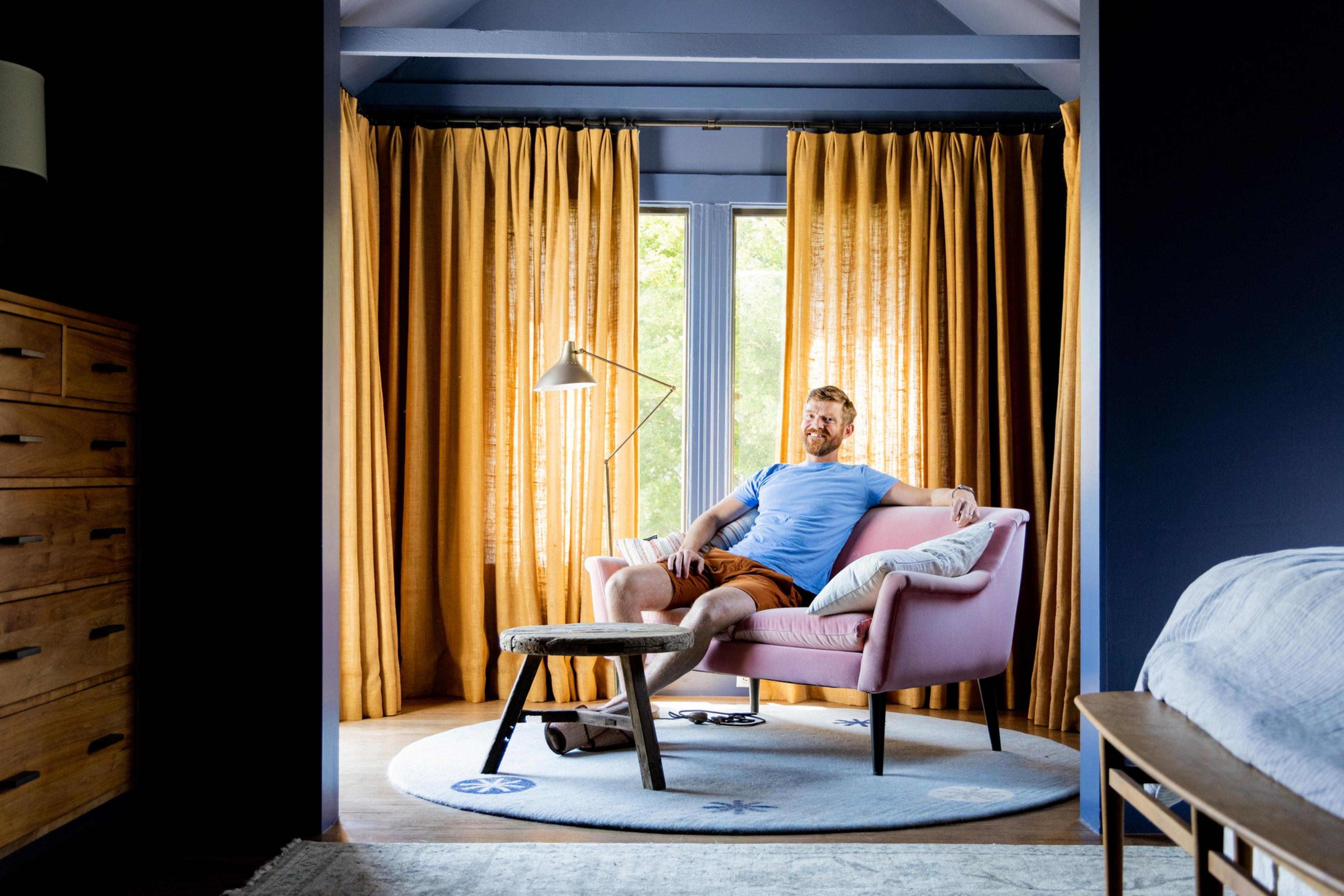 A man in a blue shirt and shorts relaxes on a pink couch in a cozy room with mustard-colored curtains. The room features a wooden dresser and a stylish floor lamp.