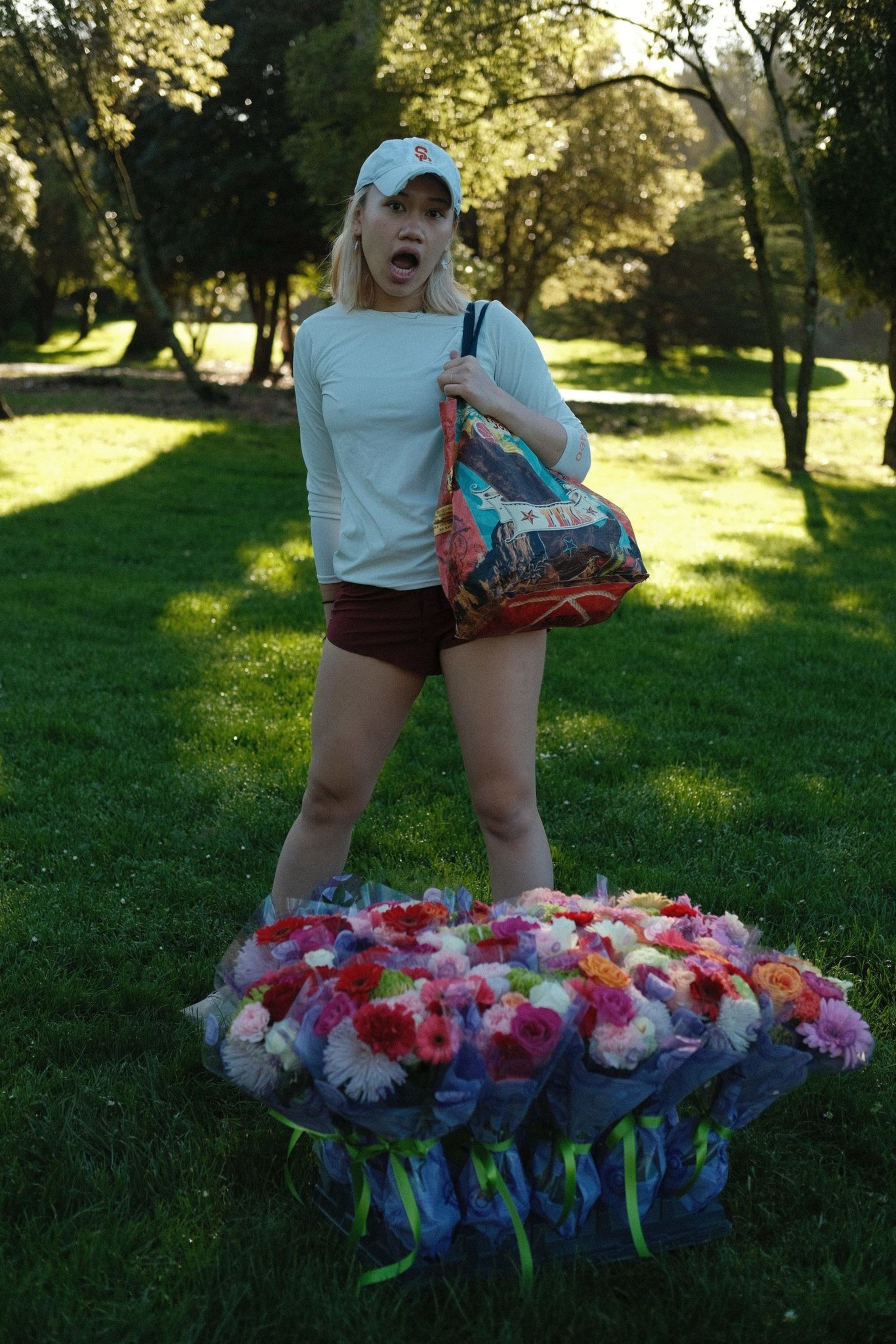A person stands on grass, wearing a cap and light top, holding a colorful bag. In front, there's a large bunch of vibrant, wrapped flowers.