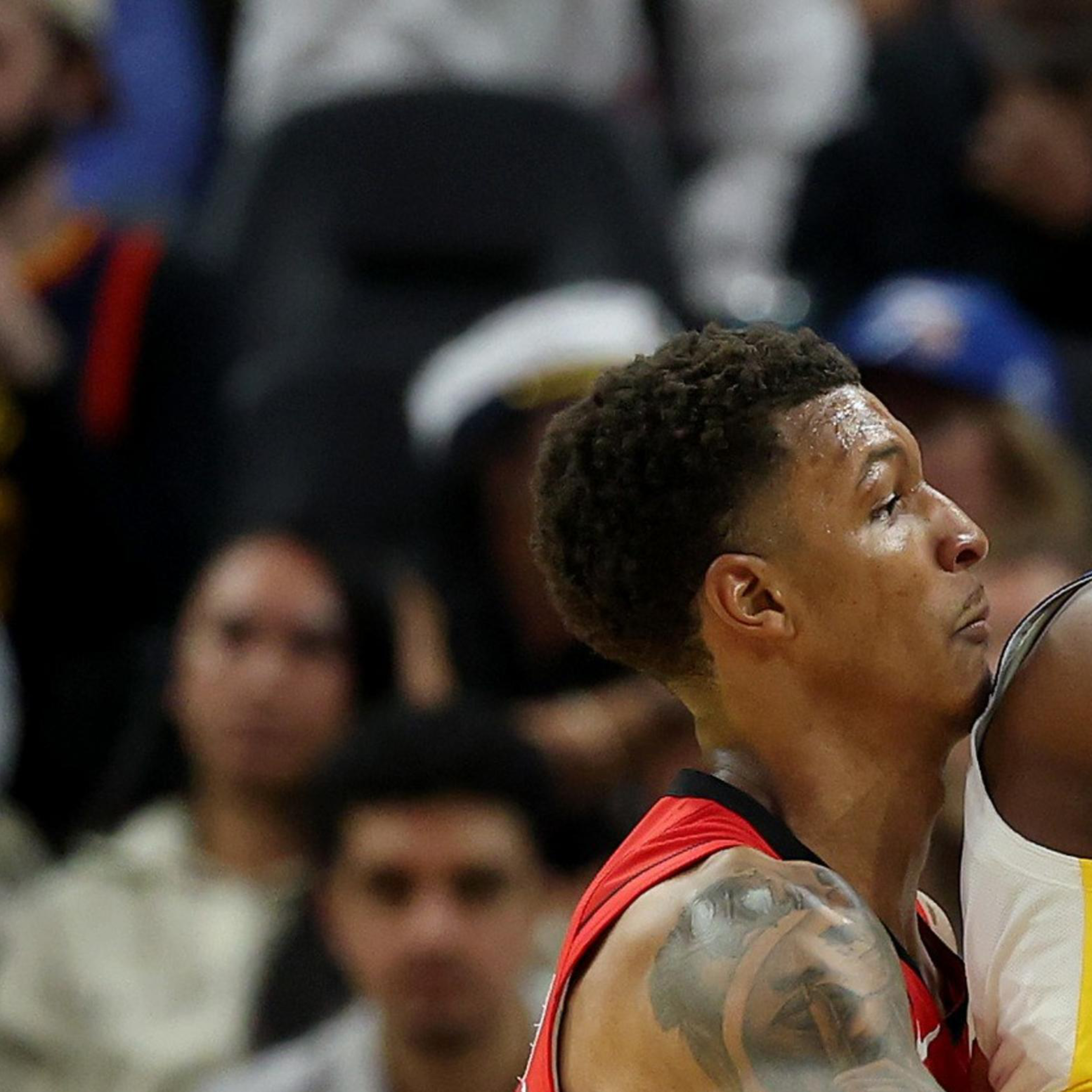 Two basketball players are in action on the court. One is wearing a red jersey, closely defending another player in a white jersey who is holding the ball. The crowd watches intently.