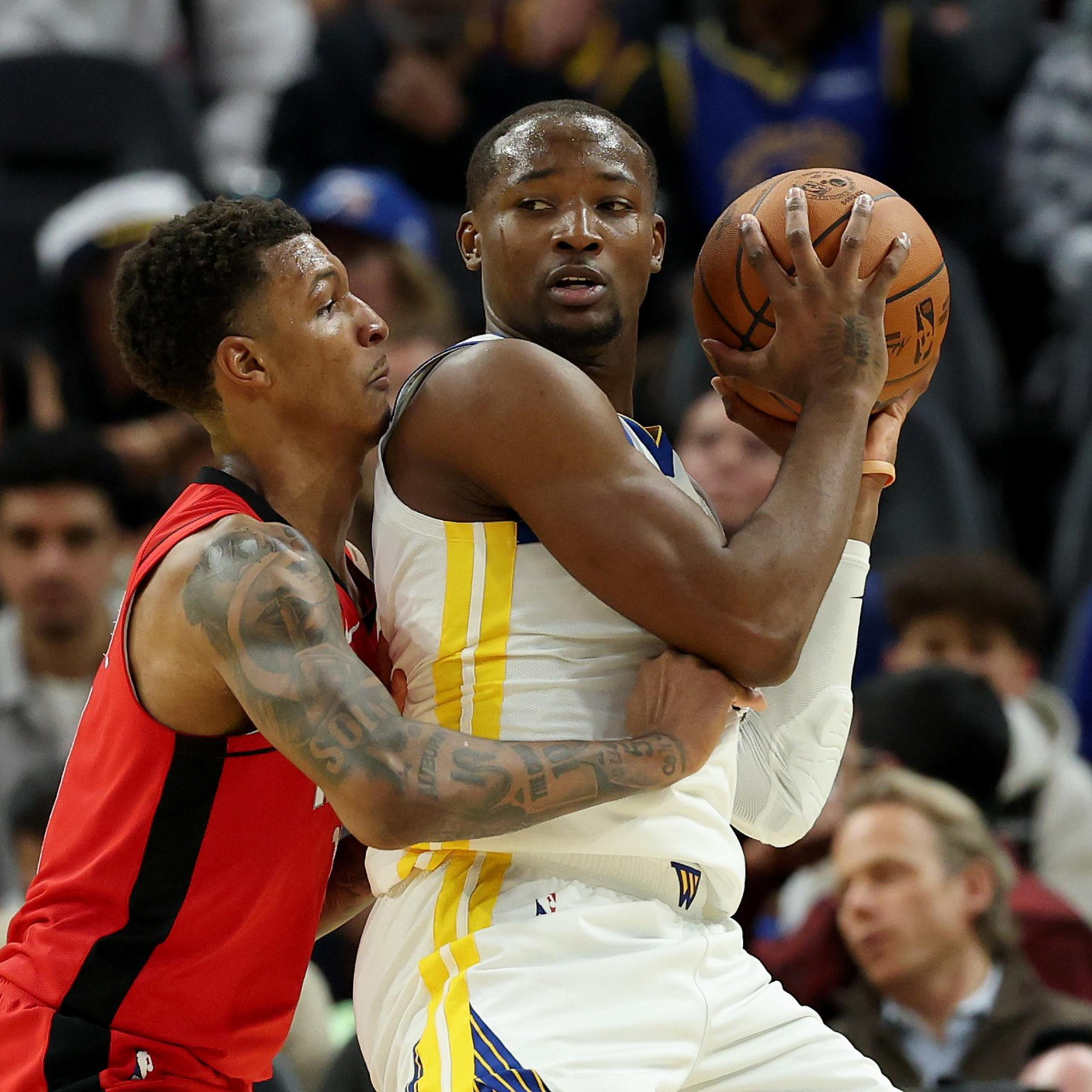 Two basketball players are in action on the court. One is wearing a red jersey, closely defending another player in a white jersey who is holding the ball. The crowd watches intently.