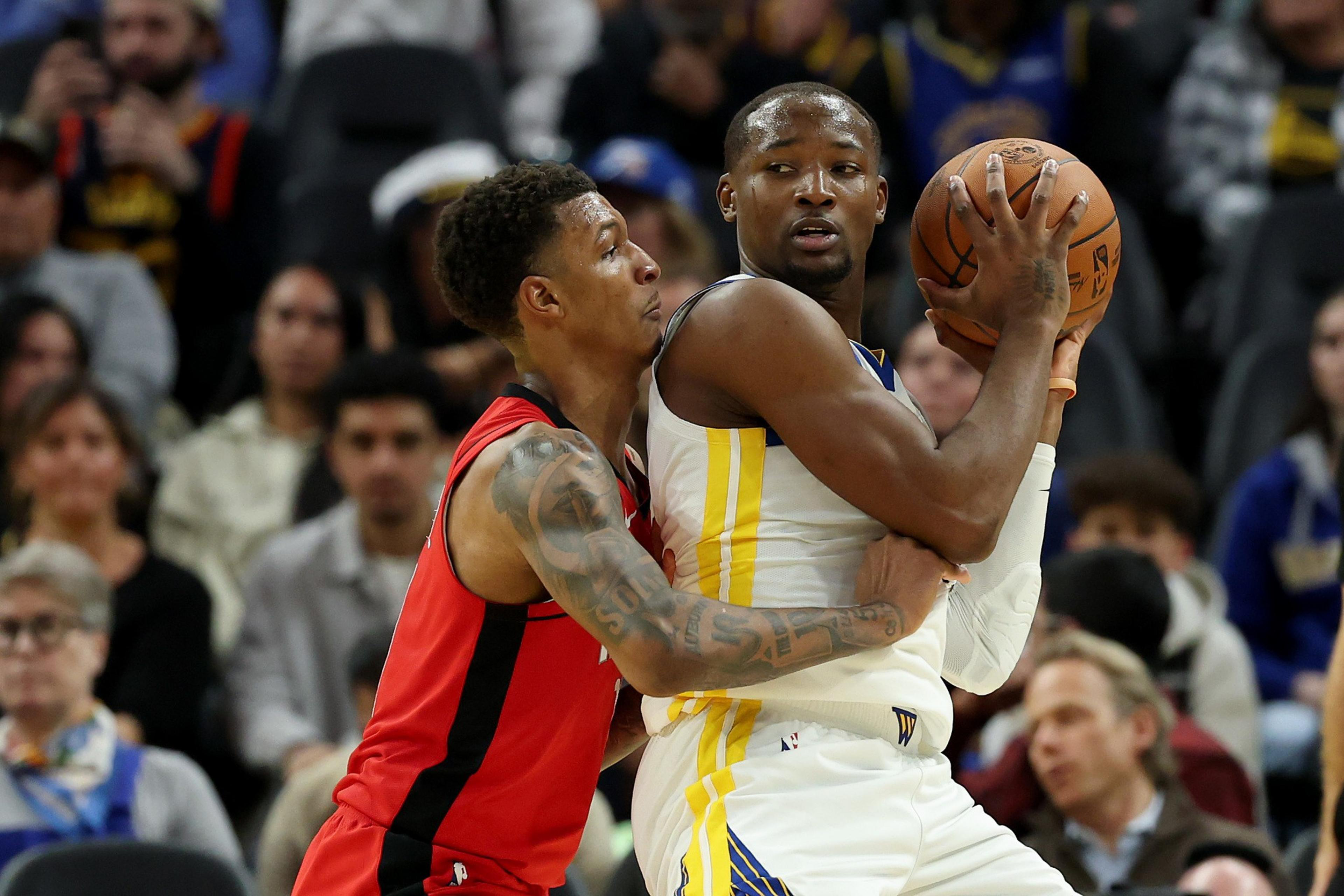 Two basketball players are in action on the court. One is wearing a red jersey, closely defending another player in a white jersey who is holding the ball. The crowd watches intently.