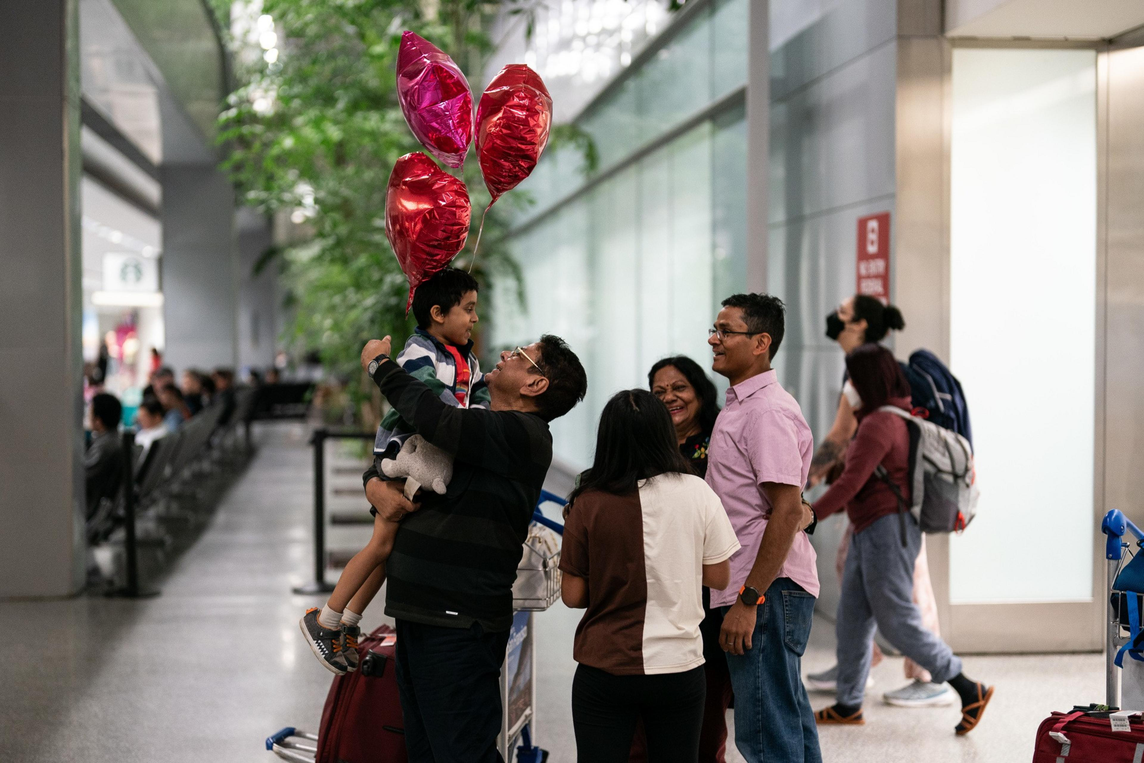 A group of people is at an airport greeting a mother and child, who is being lifted and holding red heart-shaped balloons. Luggage and travelers surround them.