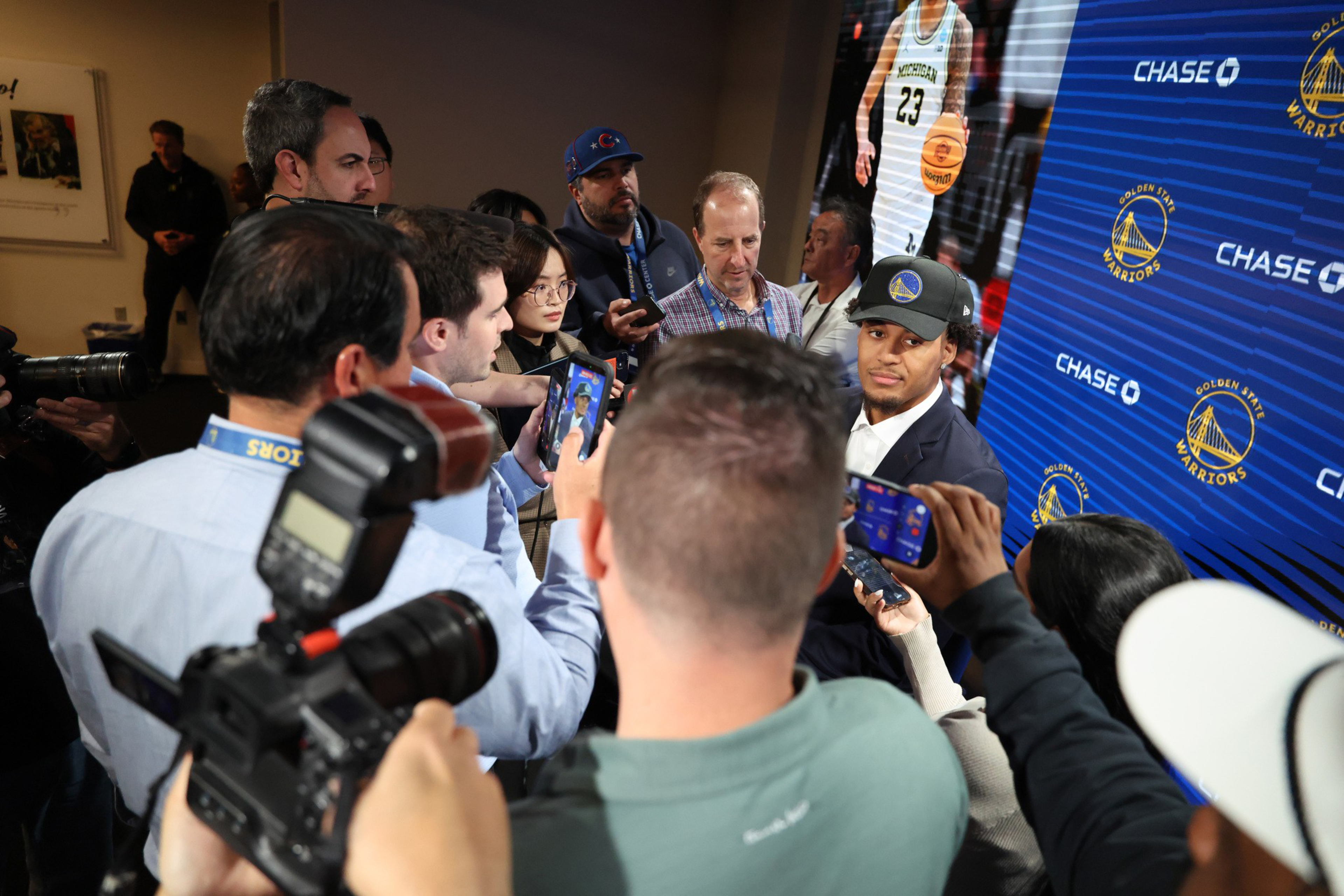 A young man wearing a Golden State Warriors cap and suit is surrounded by reporters and photographers in front of a branded backdrop.