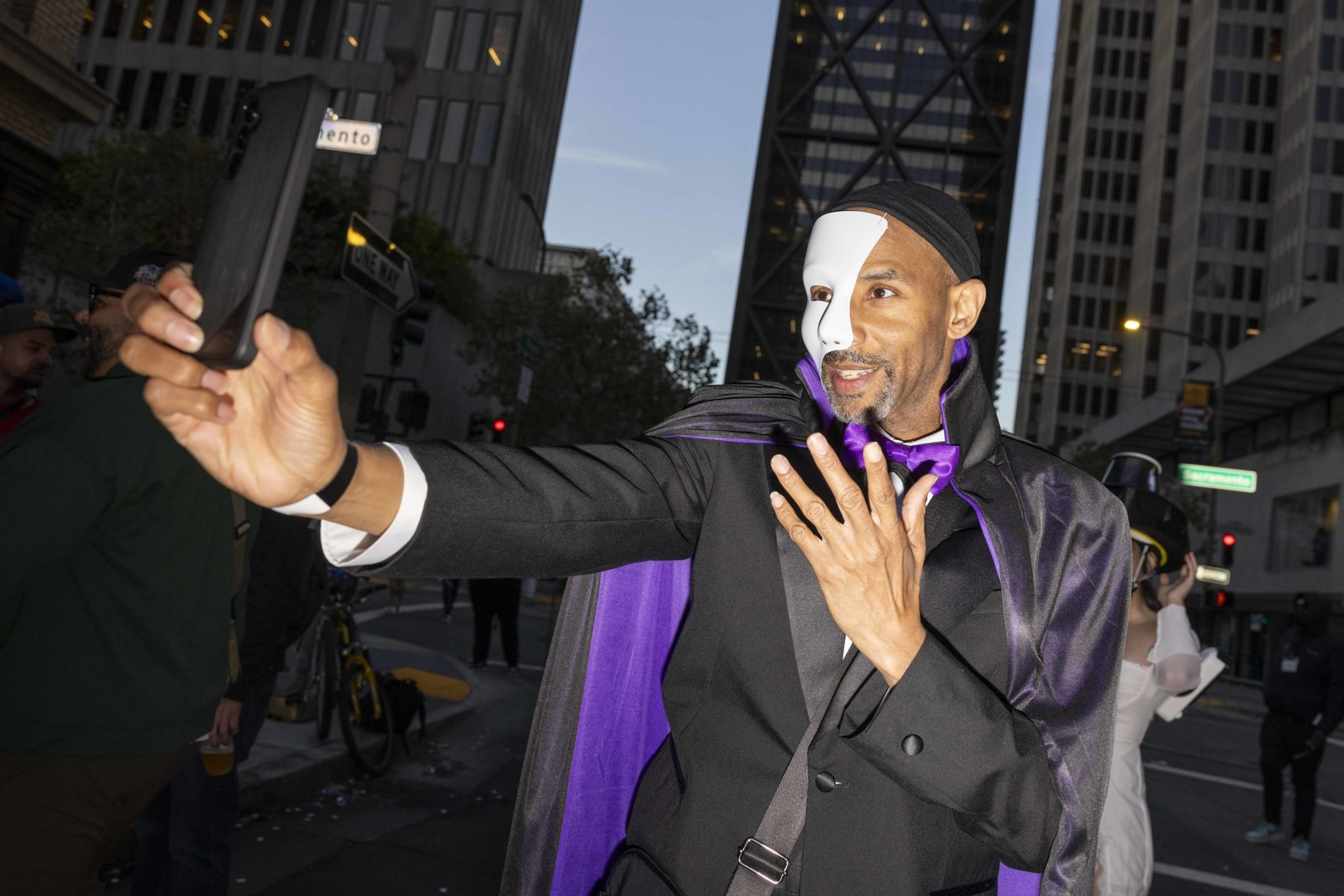 A man in a half-face white mask and black suit takes a selfie in a city street, wearing a purple cape and bow tie, with tall buildings in the background.