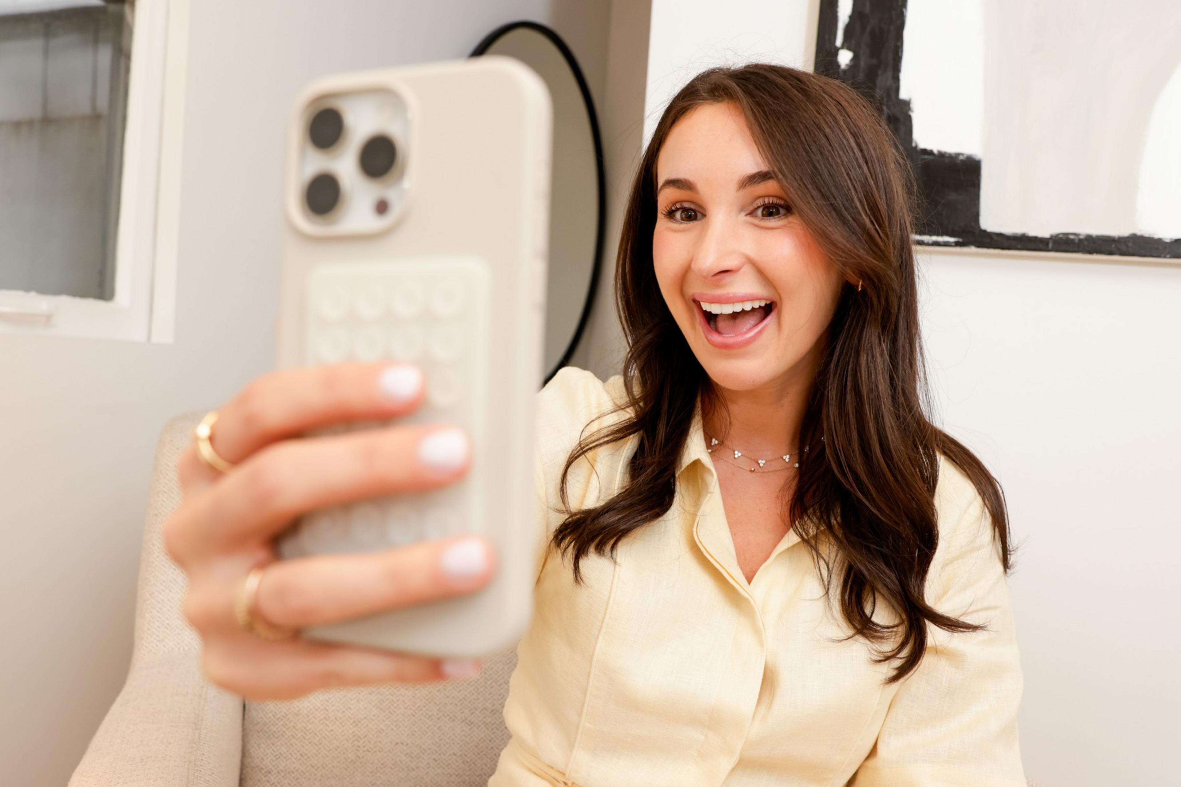 A woman with long brown hair, wearing a cream blouse, smiles widely as she takes a selfie with her white smartphone while sitting on a beige couch.