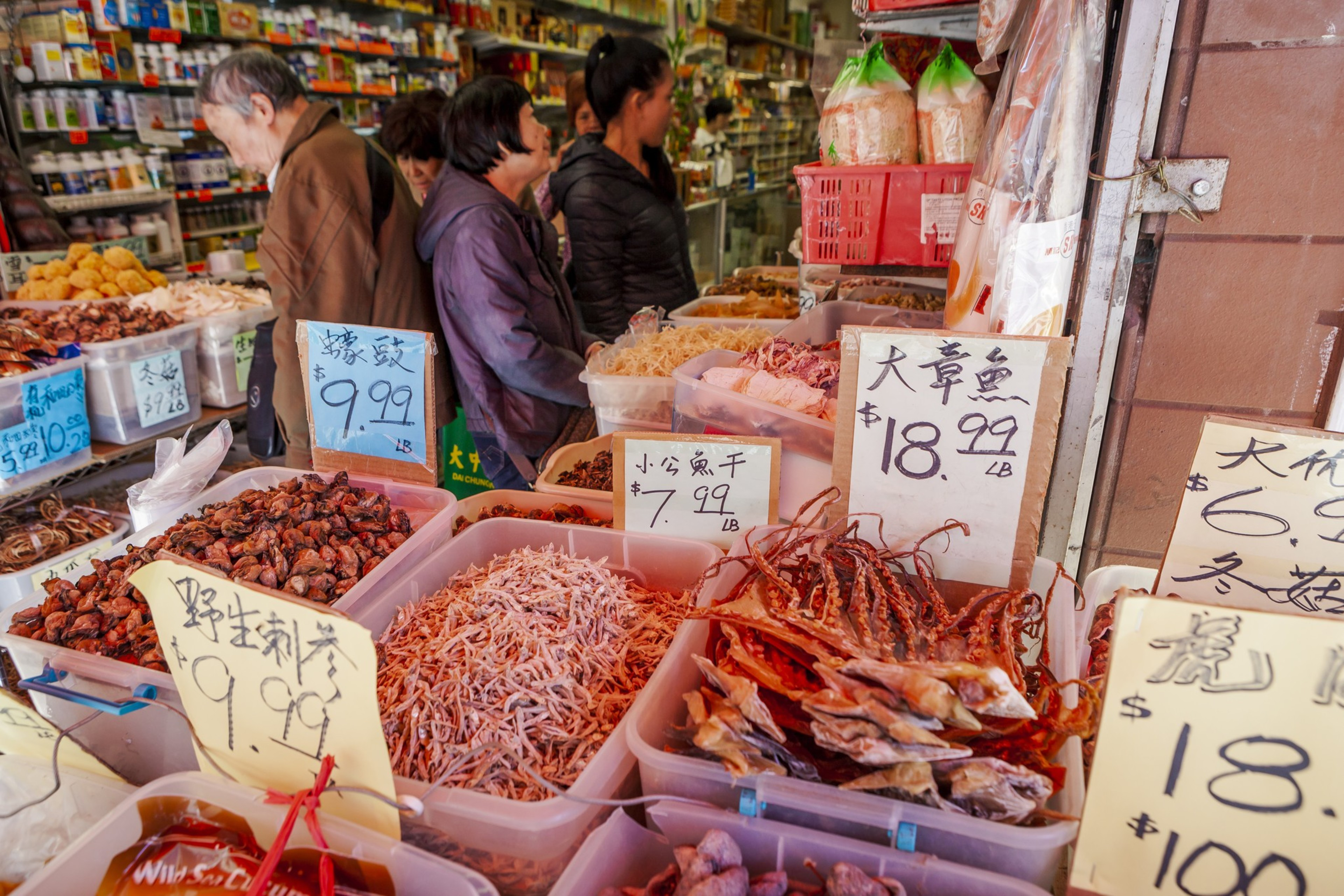 Customers at an Asian market stand browse dried seafood, herbs, and other goods in plastic bins with price tags in English and Chinese.