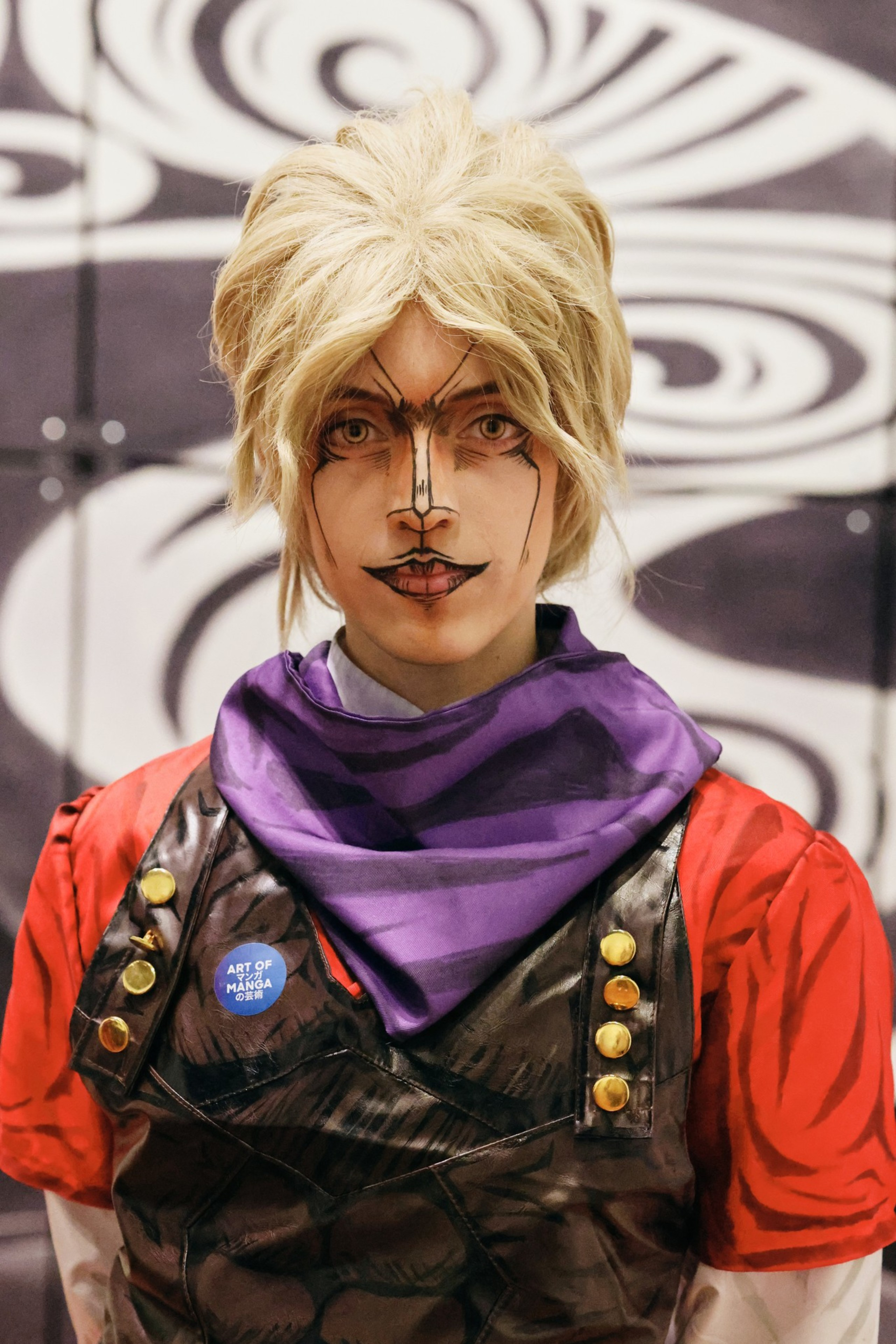 A person with blonde hair wears detailed face paint mimicking manga style, a red shirt, a purple scarf, and a vest with brass buttons and a blue “Art of Manga” badge.