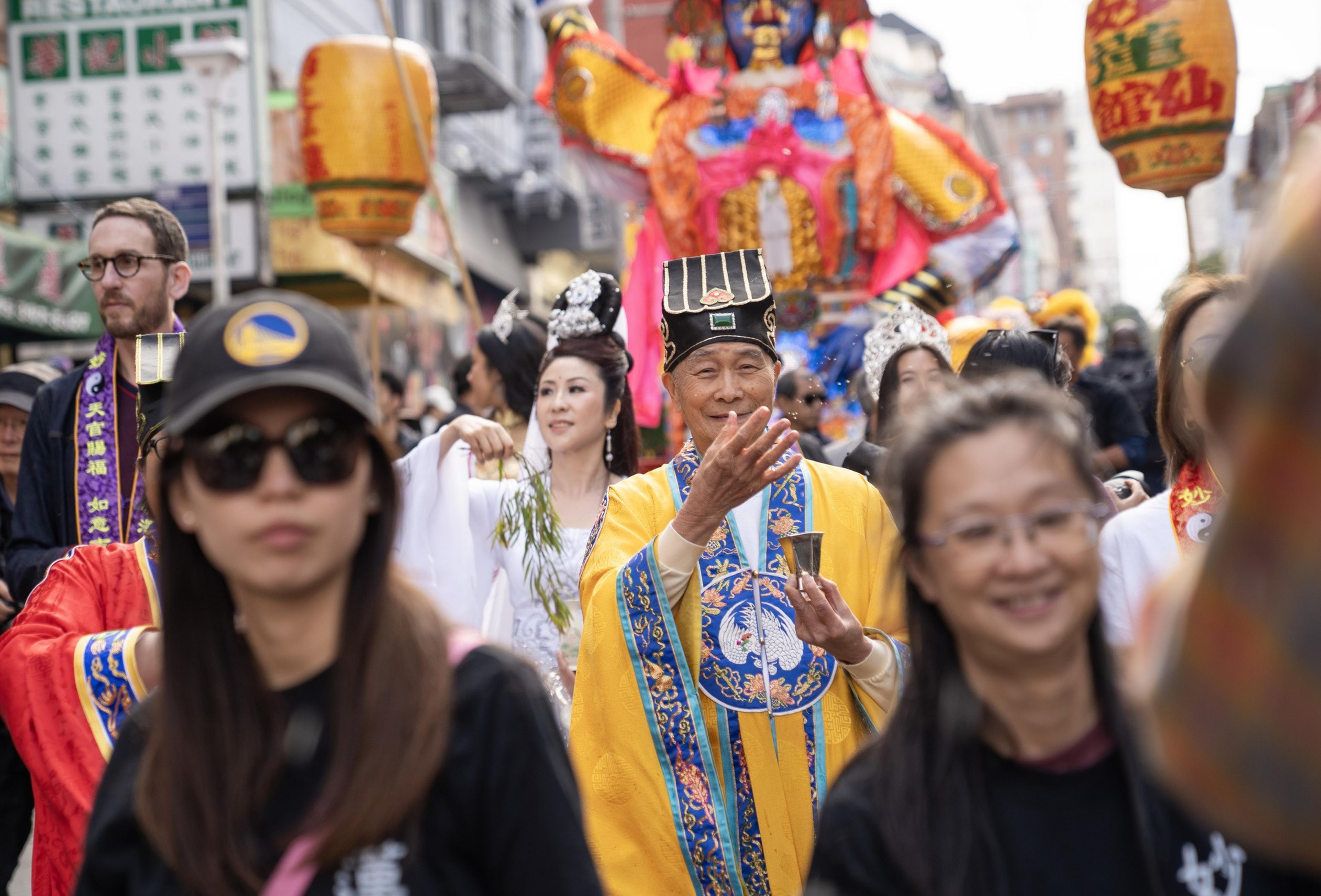 A man in a colorful traditional robe and hat holds a small cup, surrounded by people wearing festive clothes and large lanterns in a busy street parade.