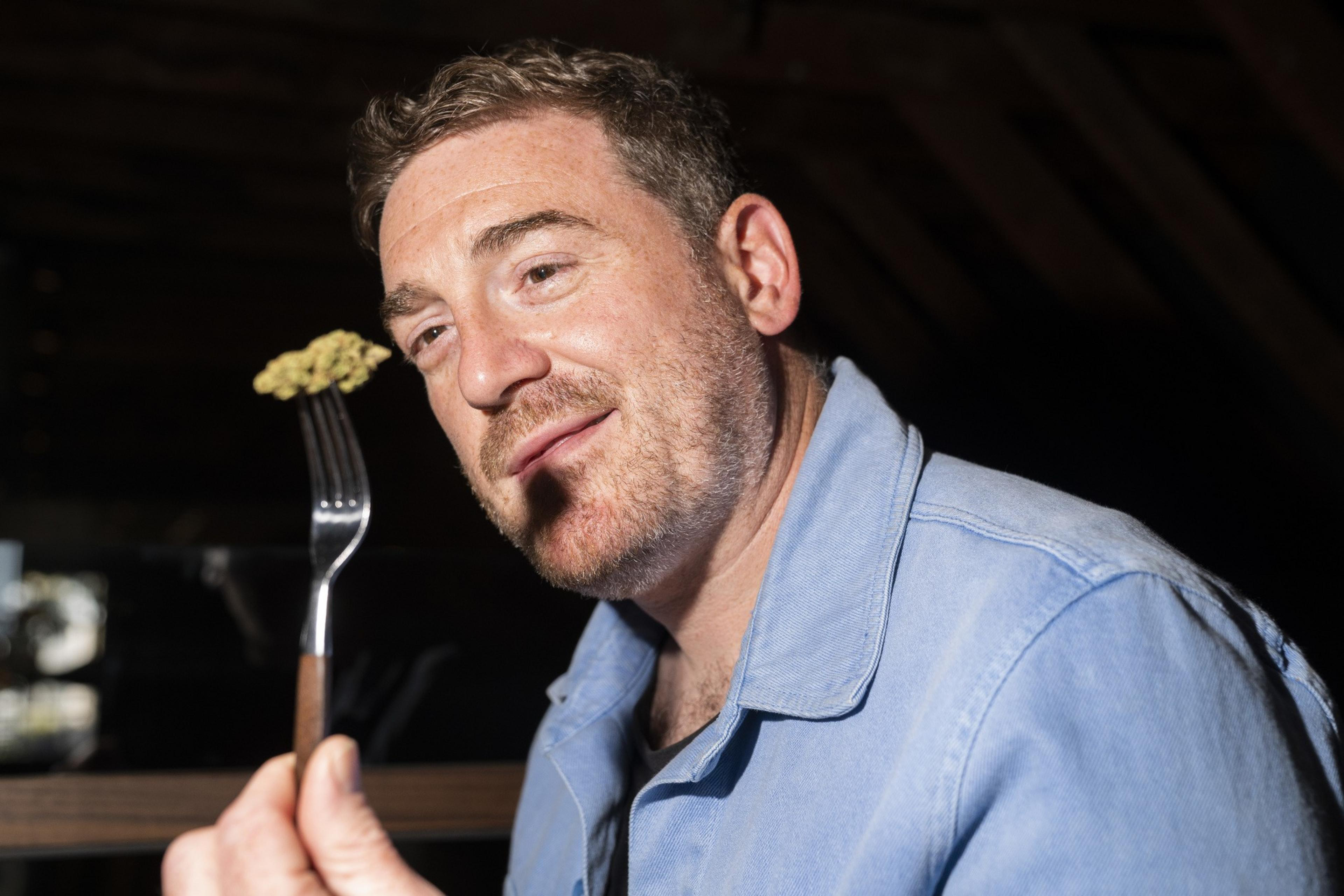 A man in a blue shirt holds up a fork with a piece of food to his face, looking at it closely. The background is dark and the lighting is focused on him.