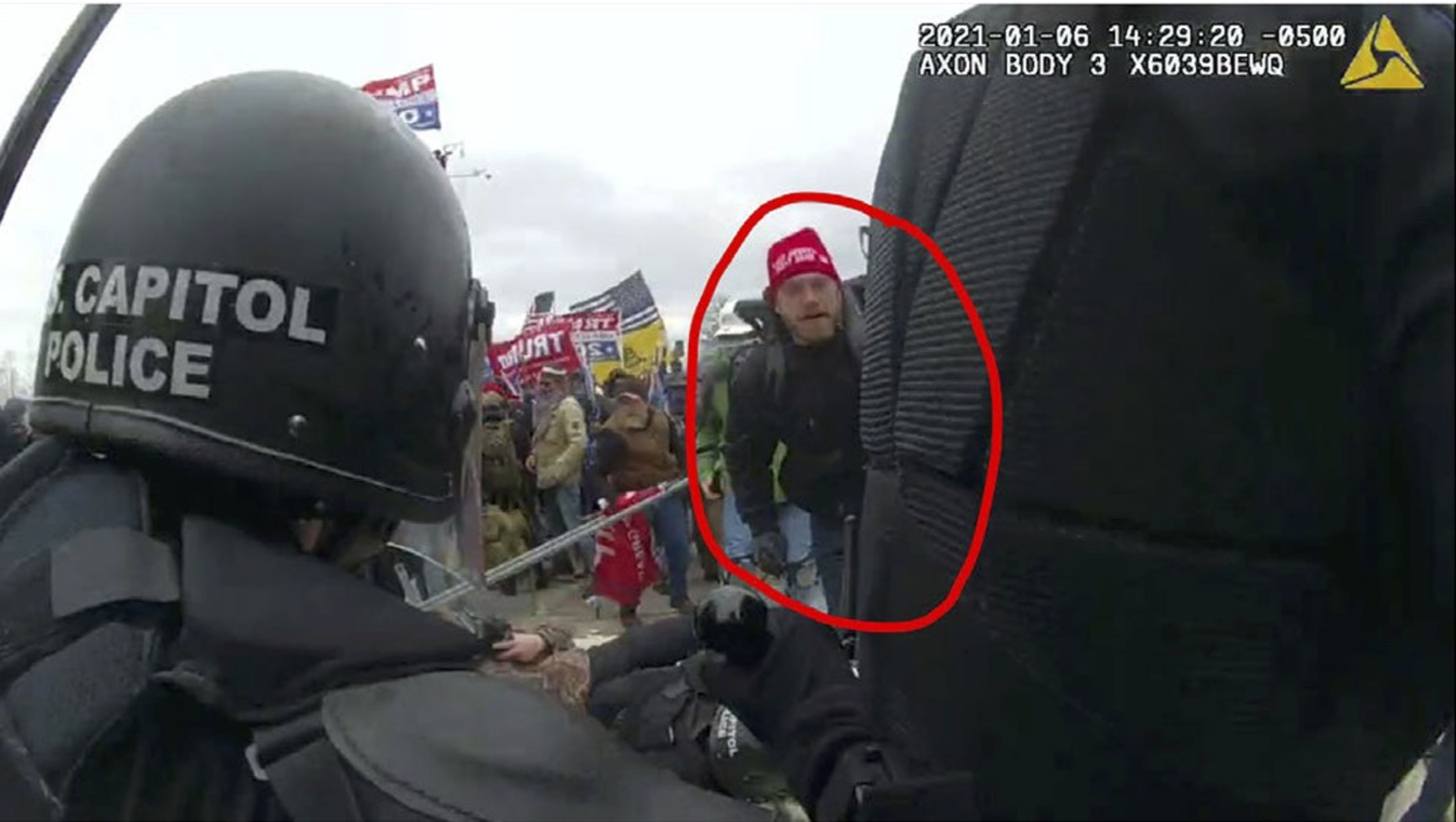 A person circled in red, wearing a red beanie, is seen in a crowd holding flags. A Capitol Police officer is in the foreground.