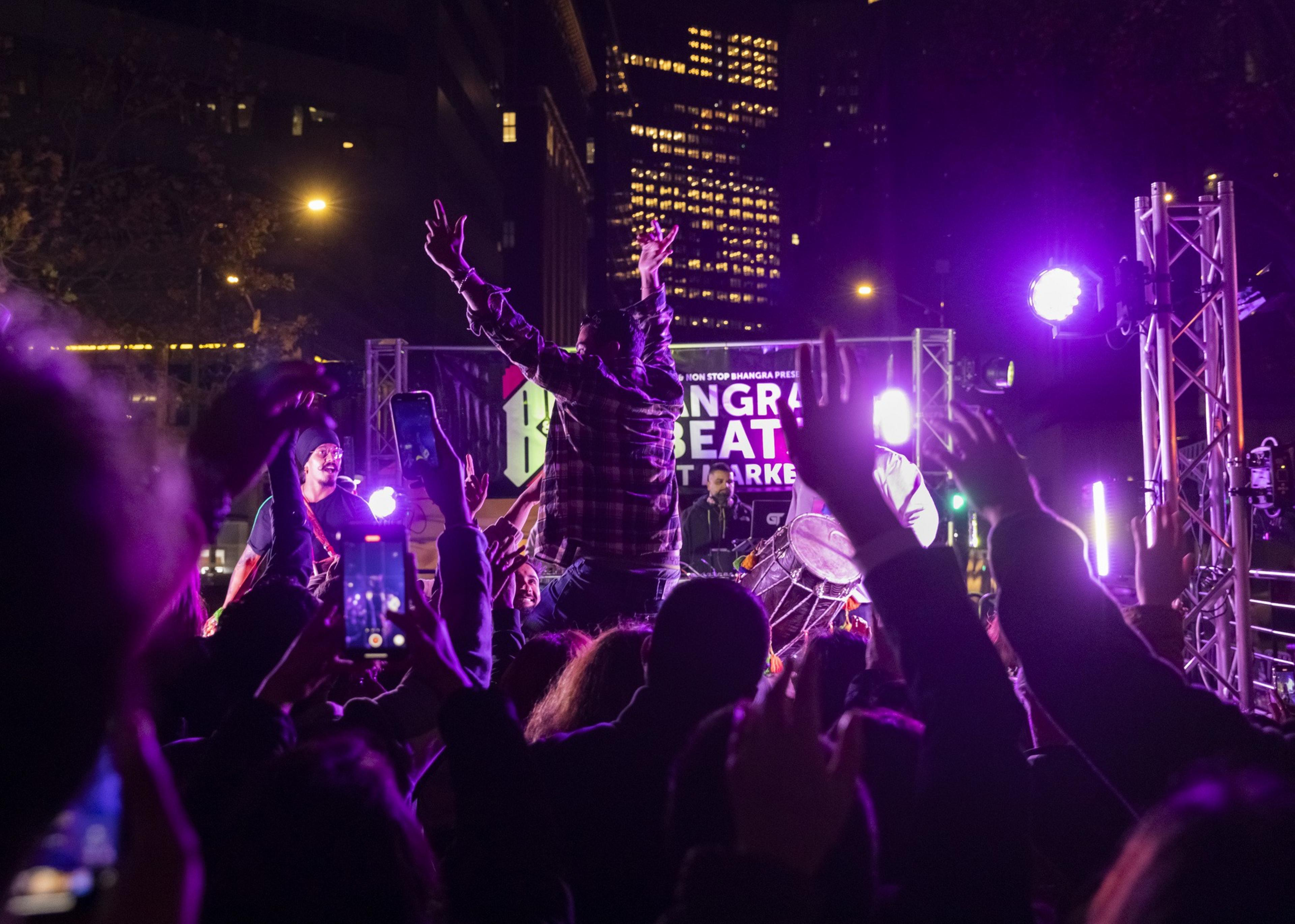 A lively night concert with people dancing and raising their hands. Bright stage lights and city buildings illuminate the scene as a performer engages with the crowd.