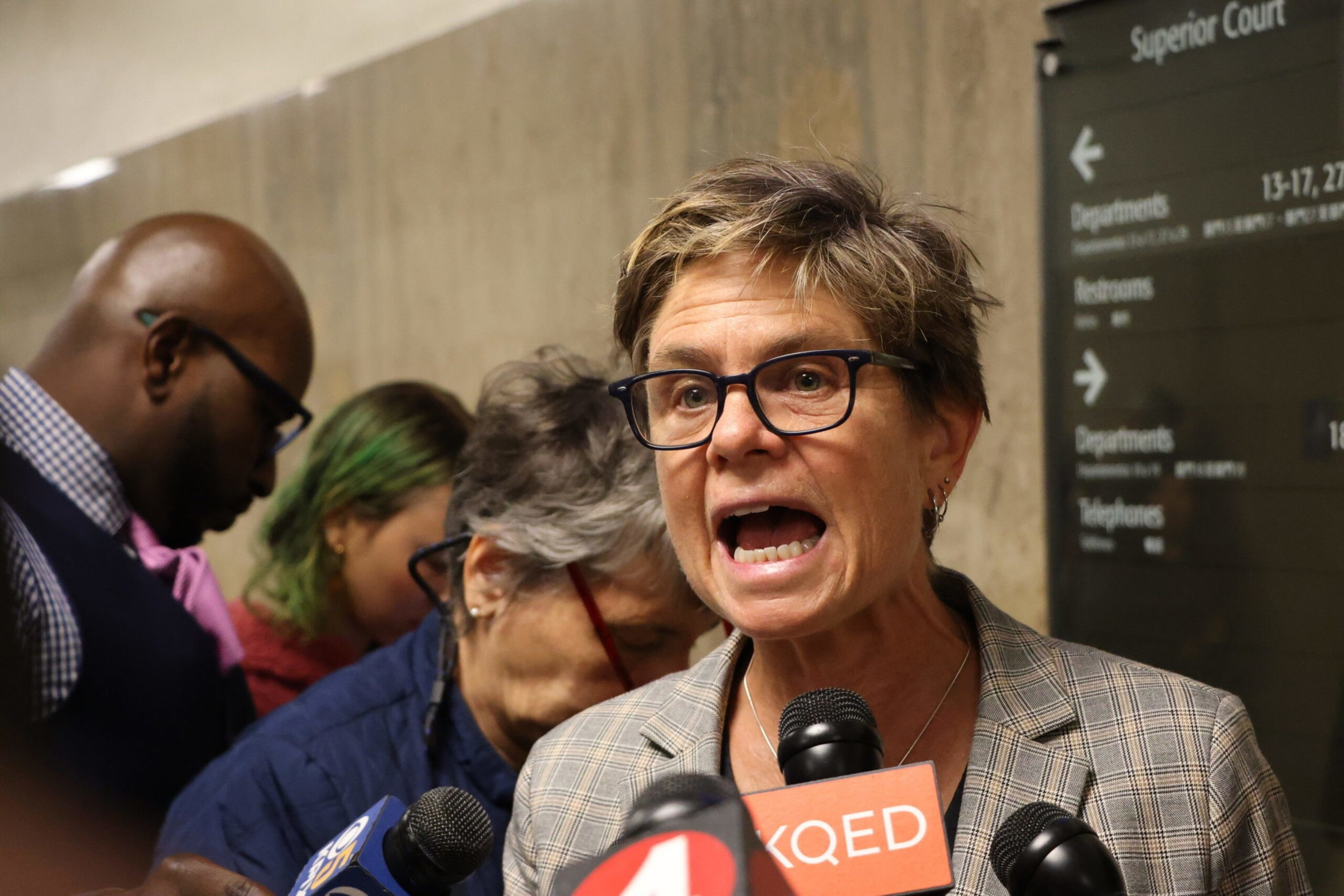 a woman in a gray suit speaks to reporters in the halllway of a courthouse.
