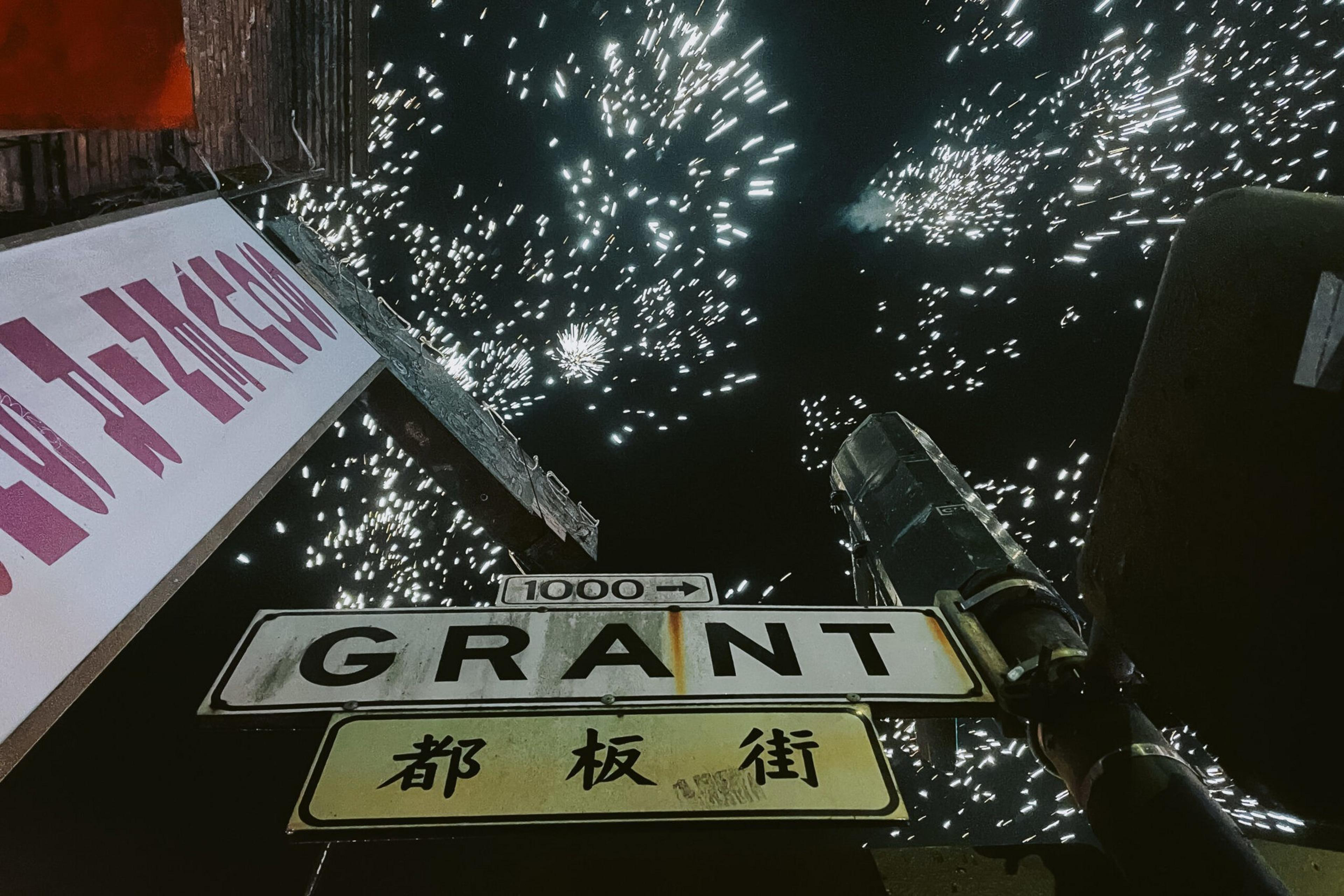 Fireworks explode brightly in the night sky above a street sign that reads "GRANT" with Chinese characters below and a "1000" address marker.
