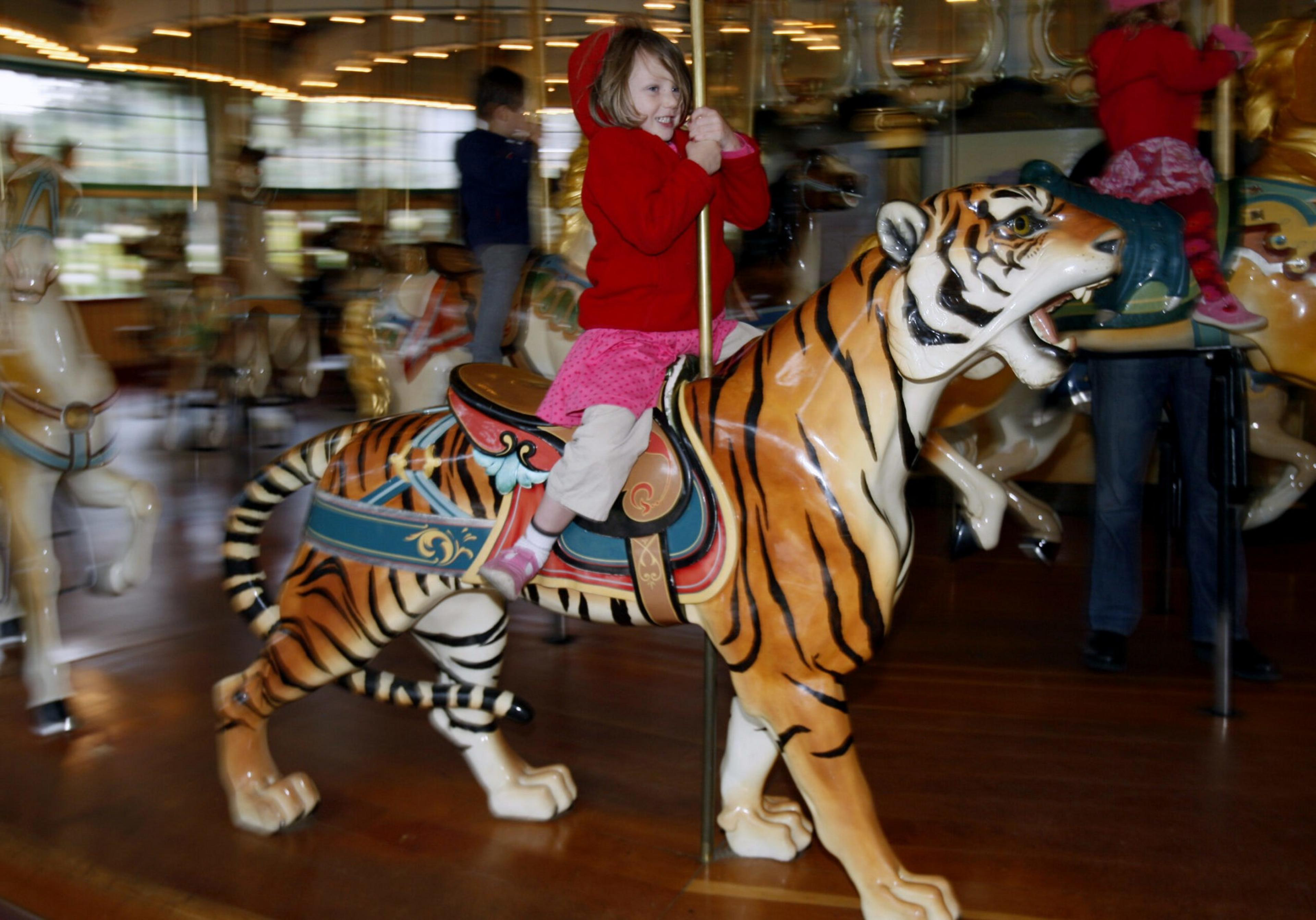 A young child in a red hoodie and pink skirt rides a carousel tiger, smiling while holding onto the pole, with blurred carousel horses in the background.