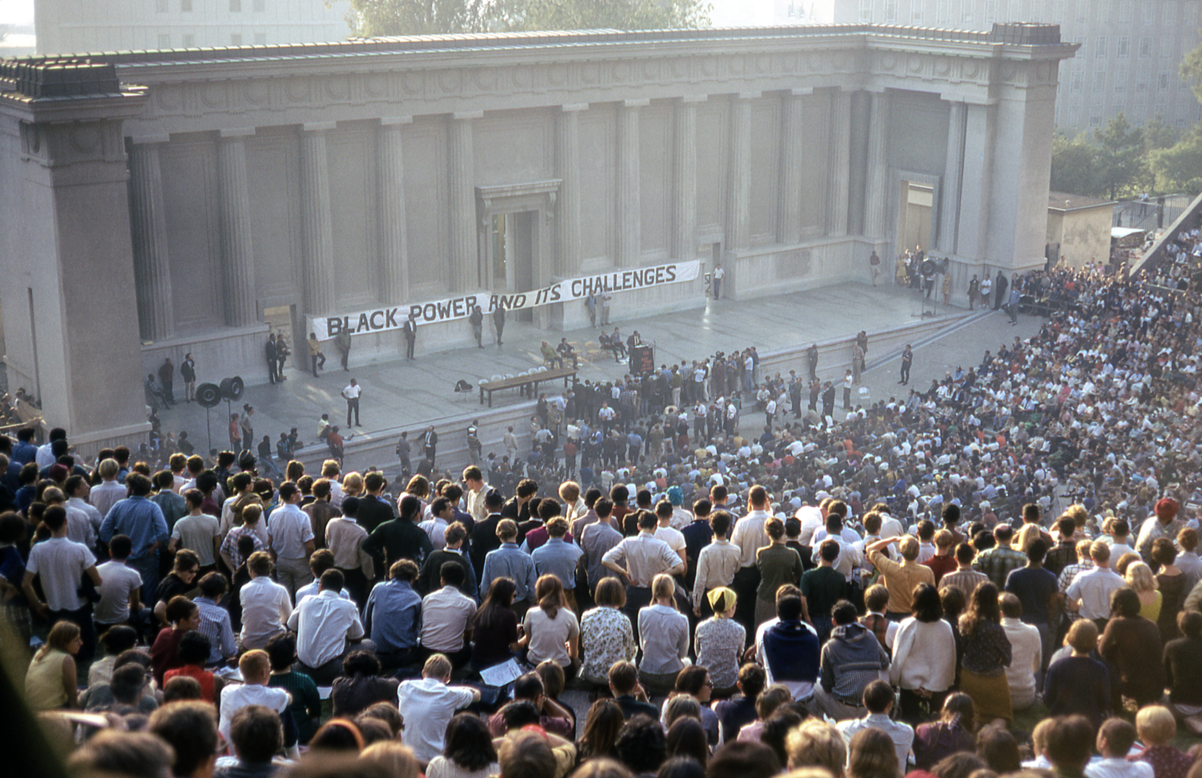 A large audience gathers in an outdoor amphitheater. A banner reads “Black Power and Its Challenges,” with speakers and people on stage.