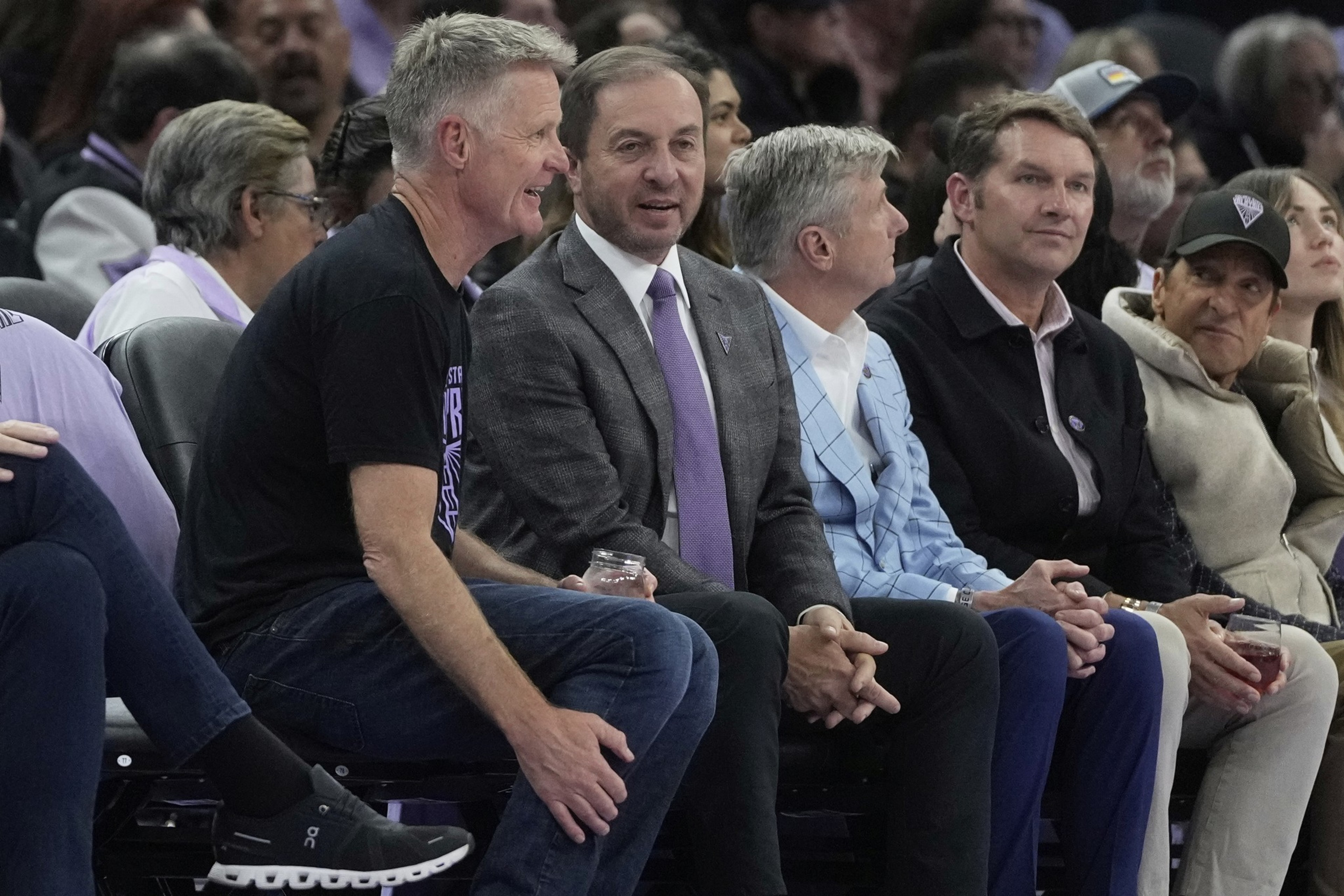 A group of men are sitting in a row at an event. One man wears a black t-shirt and jeans, laughing. Others are in suits and jackets, engaged in conversation.