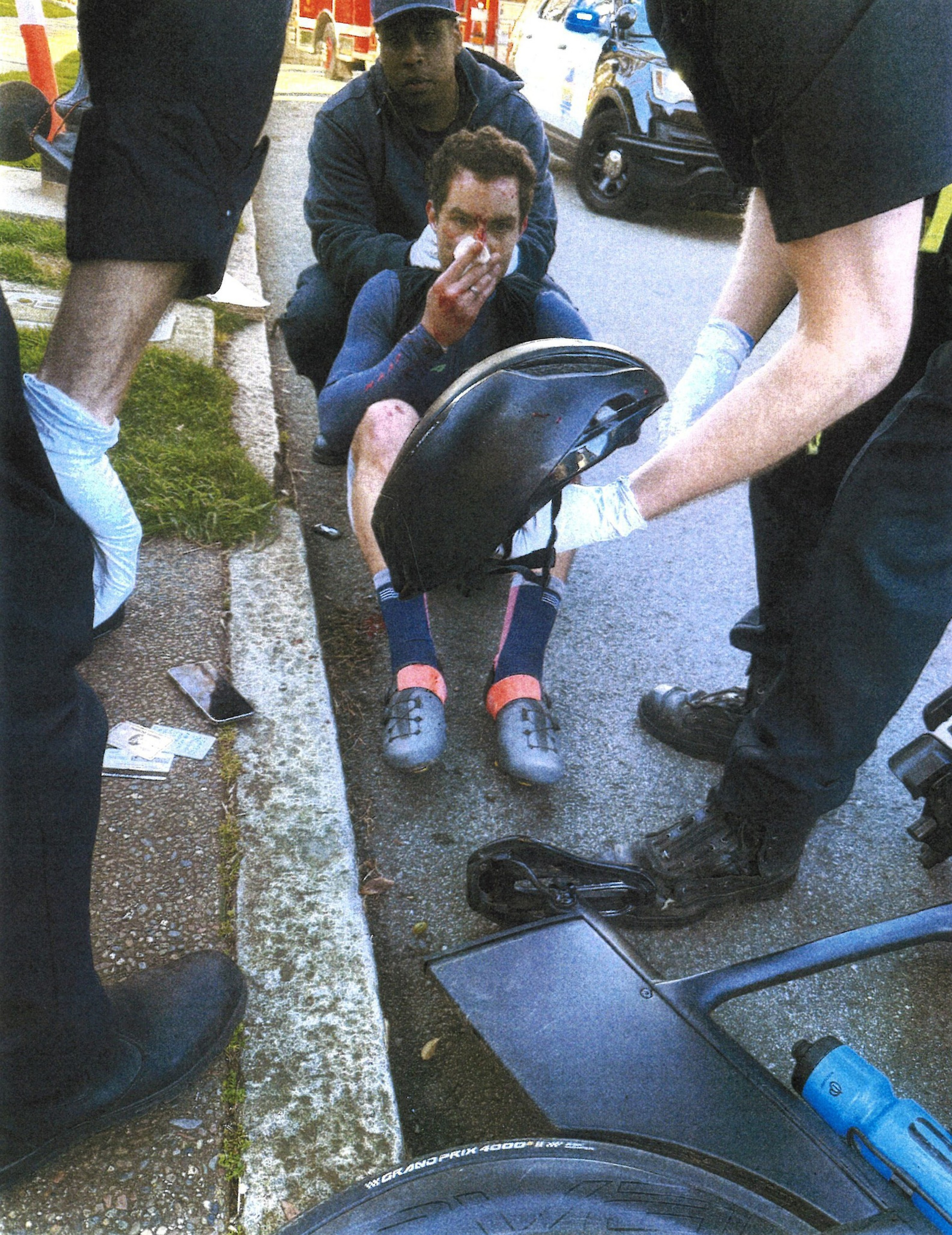 A cyclist with a bloody face sits on the curb, holding a helmet, surrounded by responders wearing gloves, with a police car and a fire truck in the background.