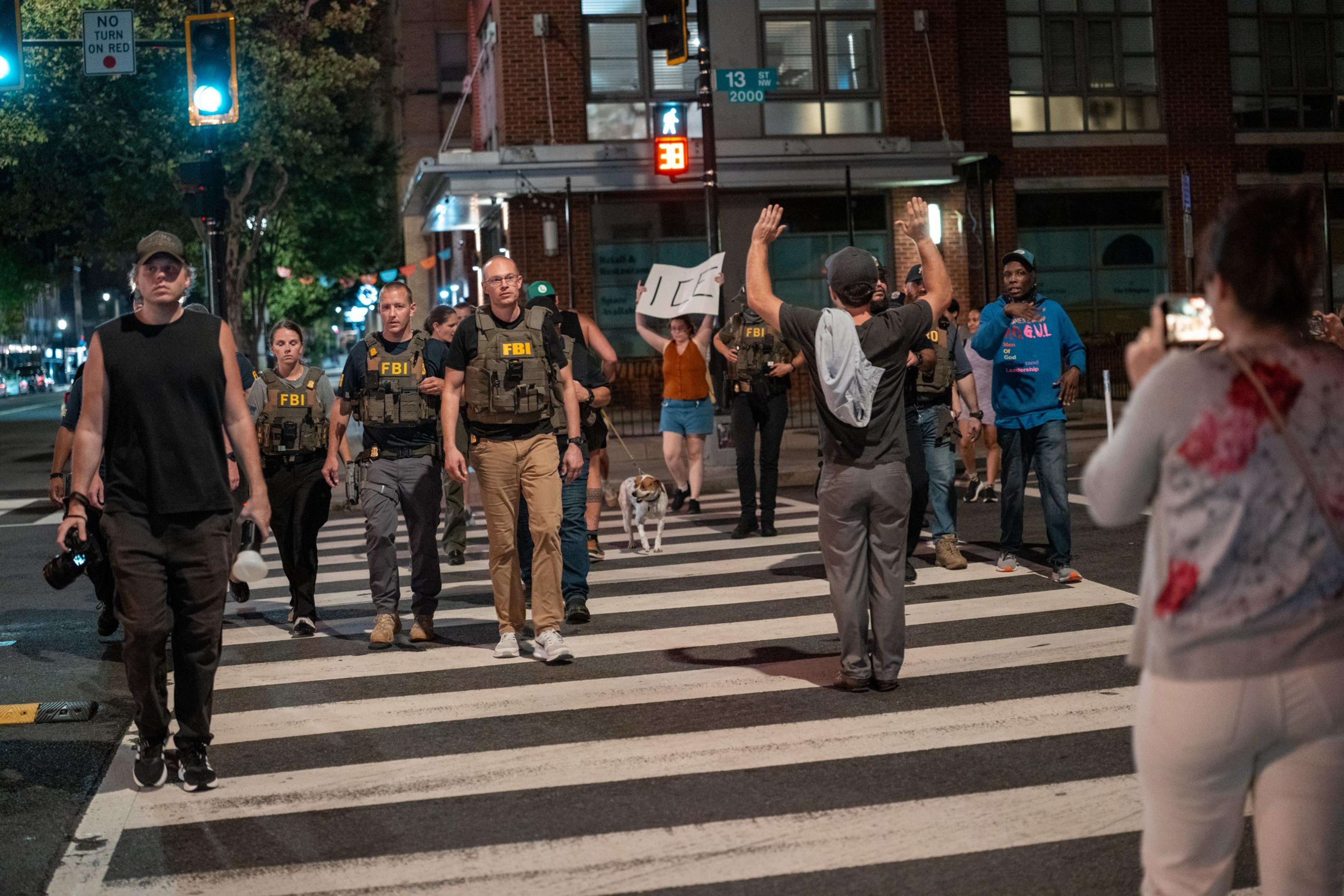 A group of FBI agents and civilians cross a street at night while a person raises hands and another takes a photo nearby.