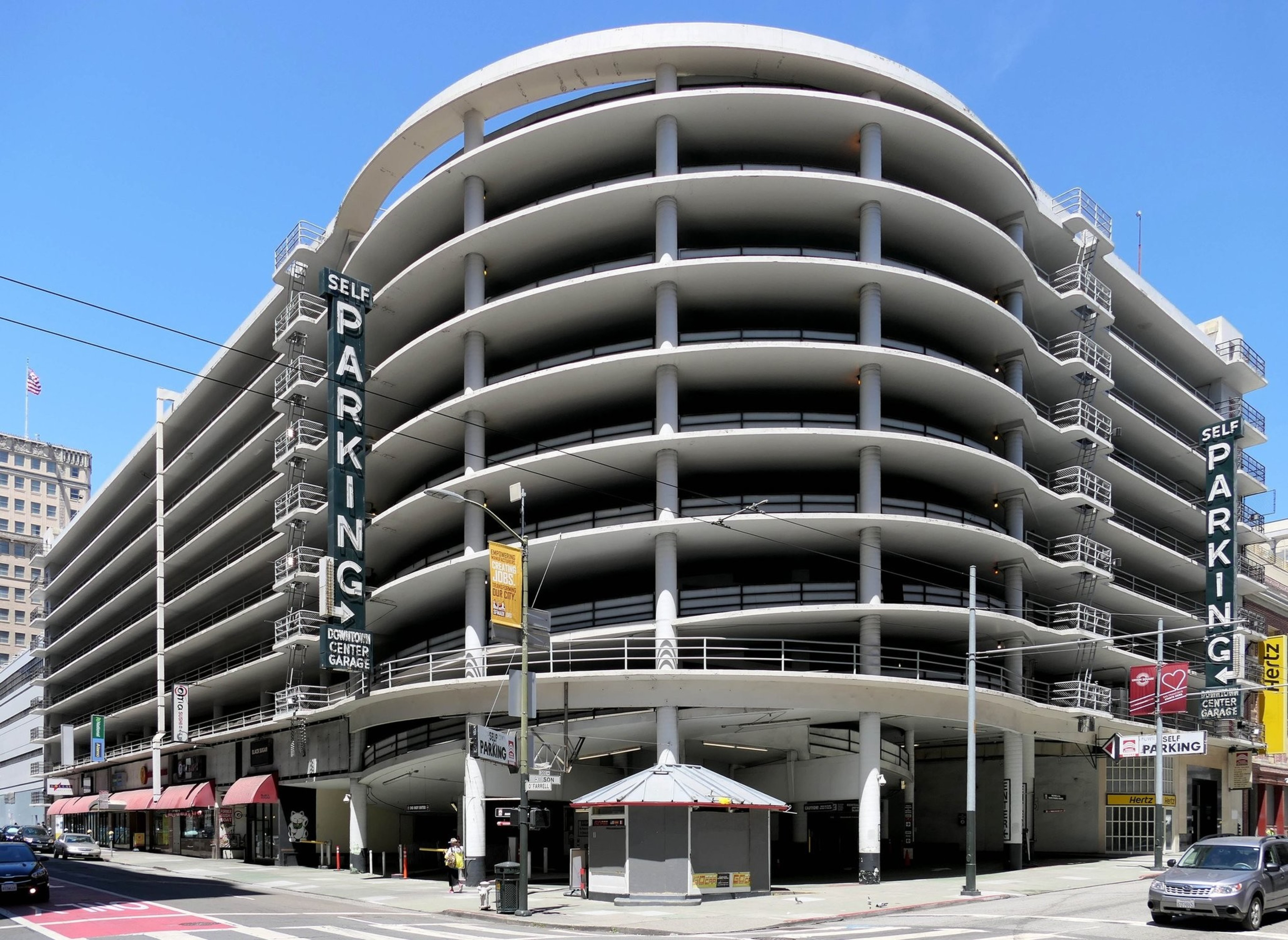 The image shows a multi-story, circular parking garage with signs saying &quot;Self Parking.&quot; It has open sides and storefronts at street level.