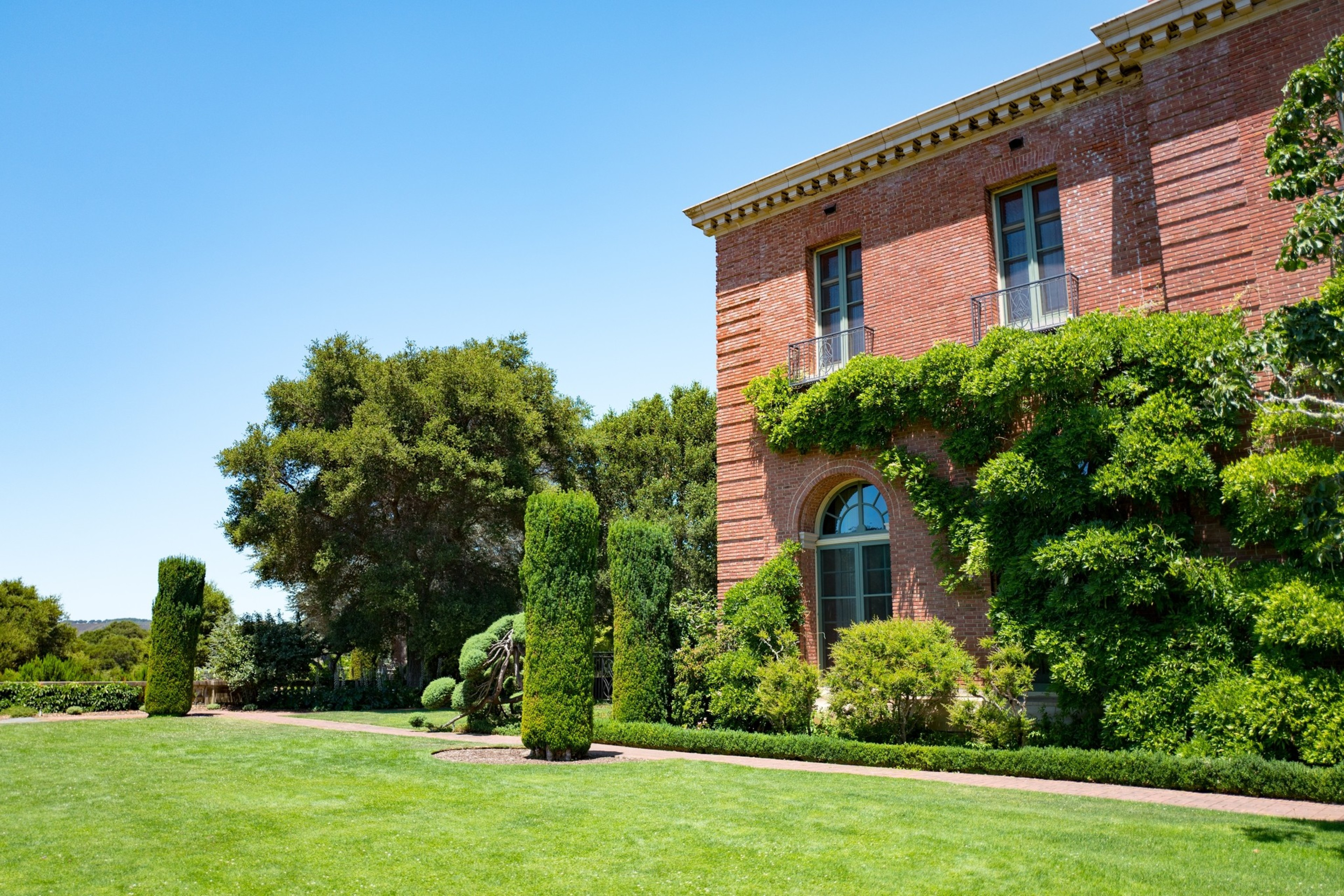 A brick building with ivy growing on its exterior and to manicured lawn, including hedges and lush trees.