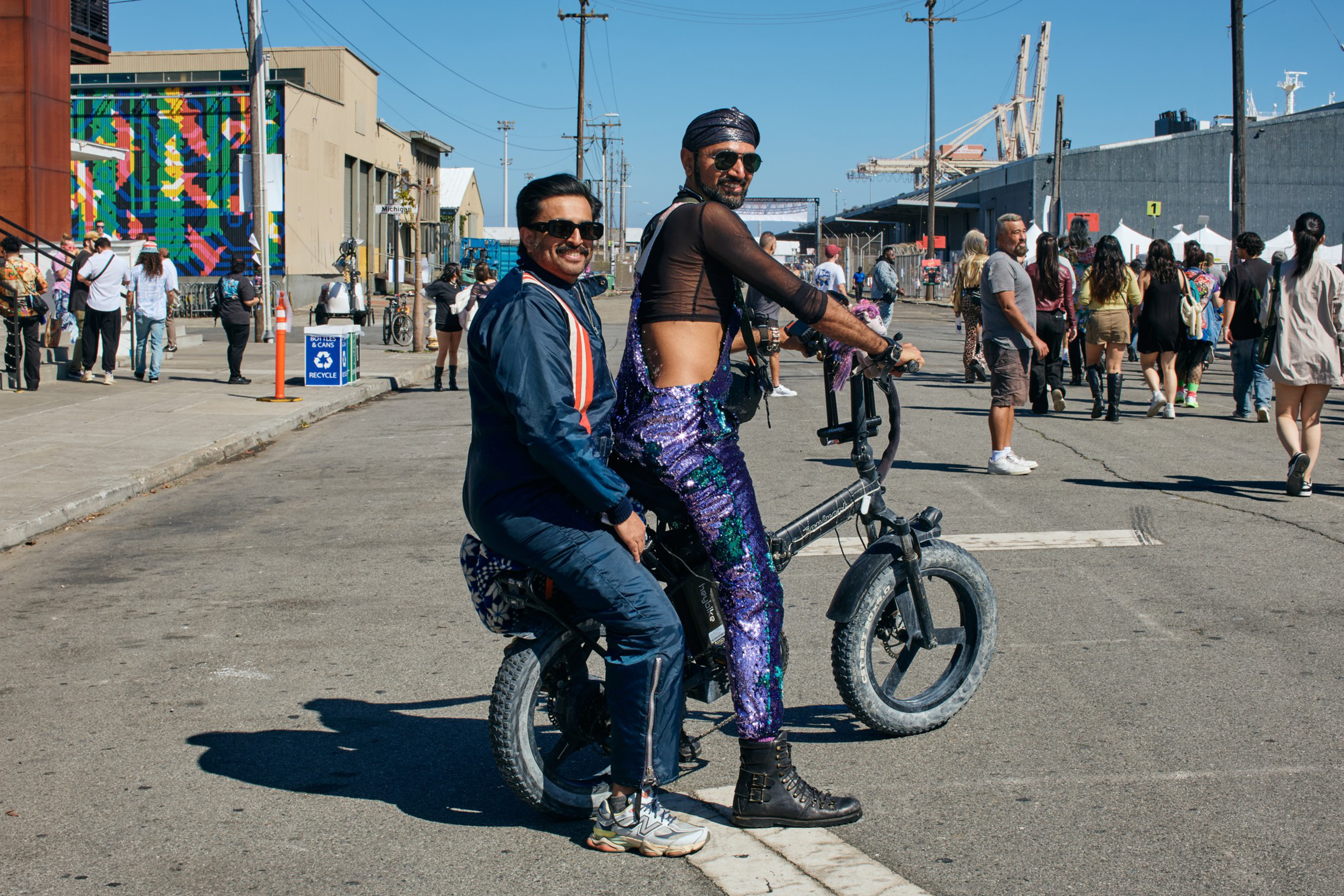 Two men are on a small bike; one wears a dark tracksuit and sunglasses, the other sparkly purple pants with a black mesh top and sunglasses. People walk in the background.